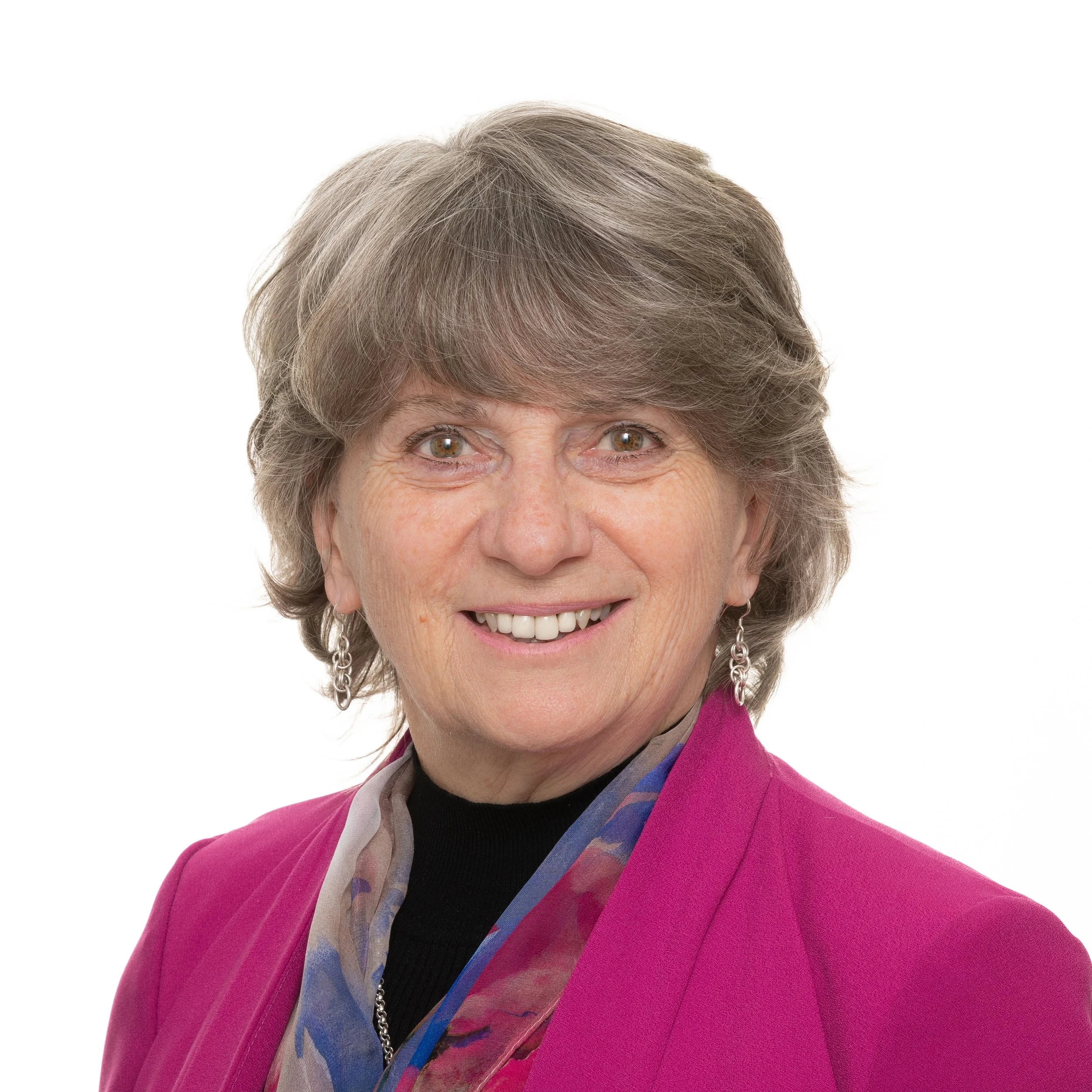 A portrait of a smiling woman with gray hair, wearing a pink blazer, a multicolored scarf, and dangling earrings, against a white background.