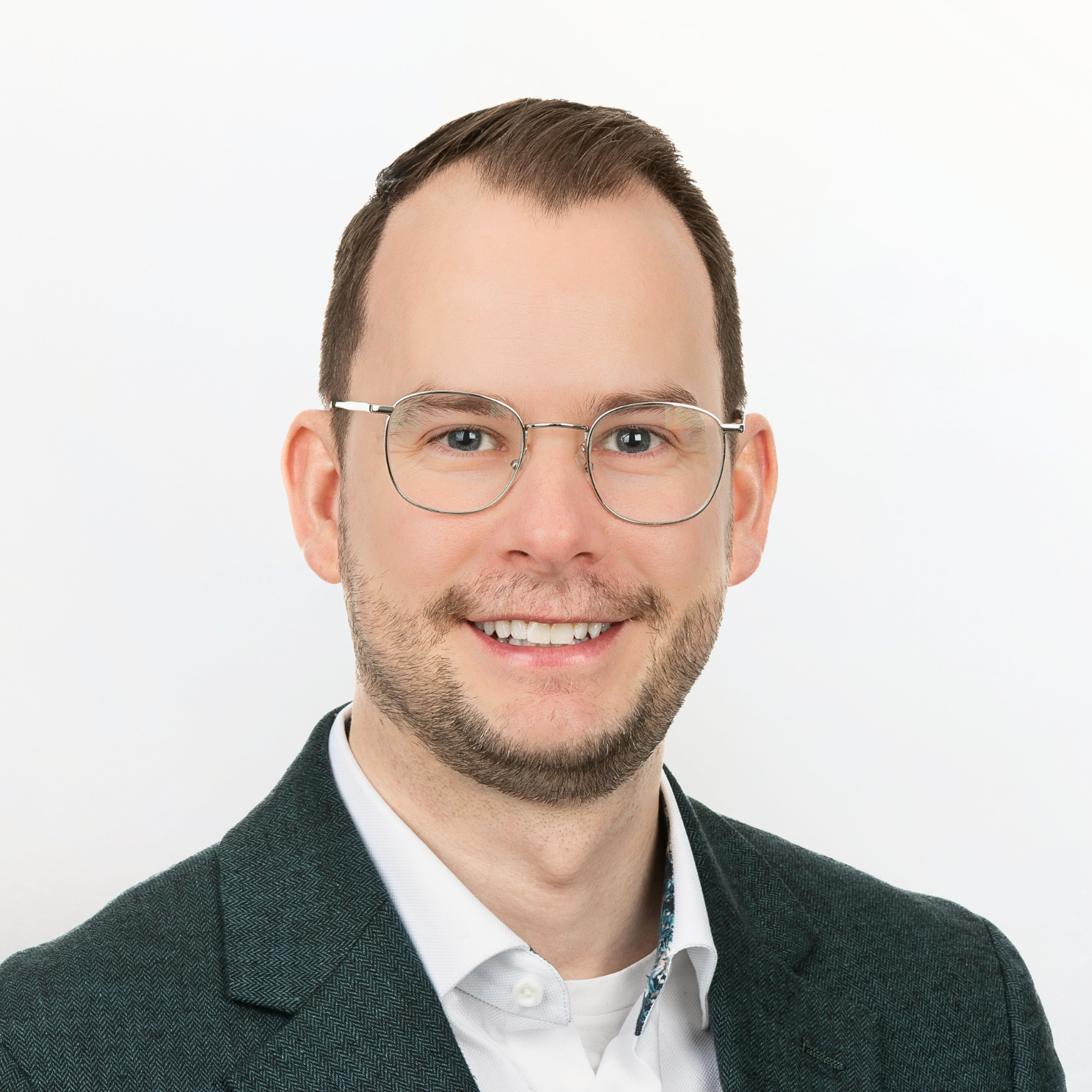 Professional headshot of a smiling man wearing glasses, a dark blazer, and a white shirt with a patterned collar, against a plain white background.