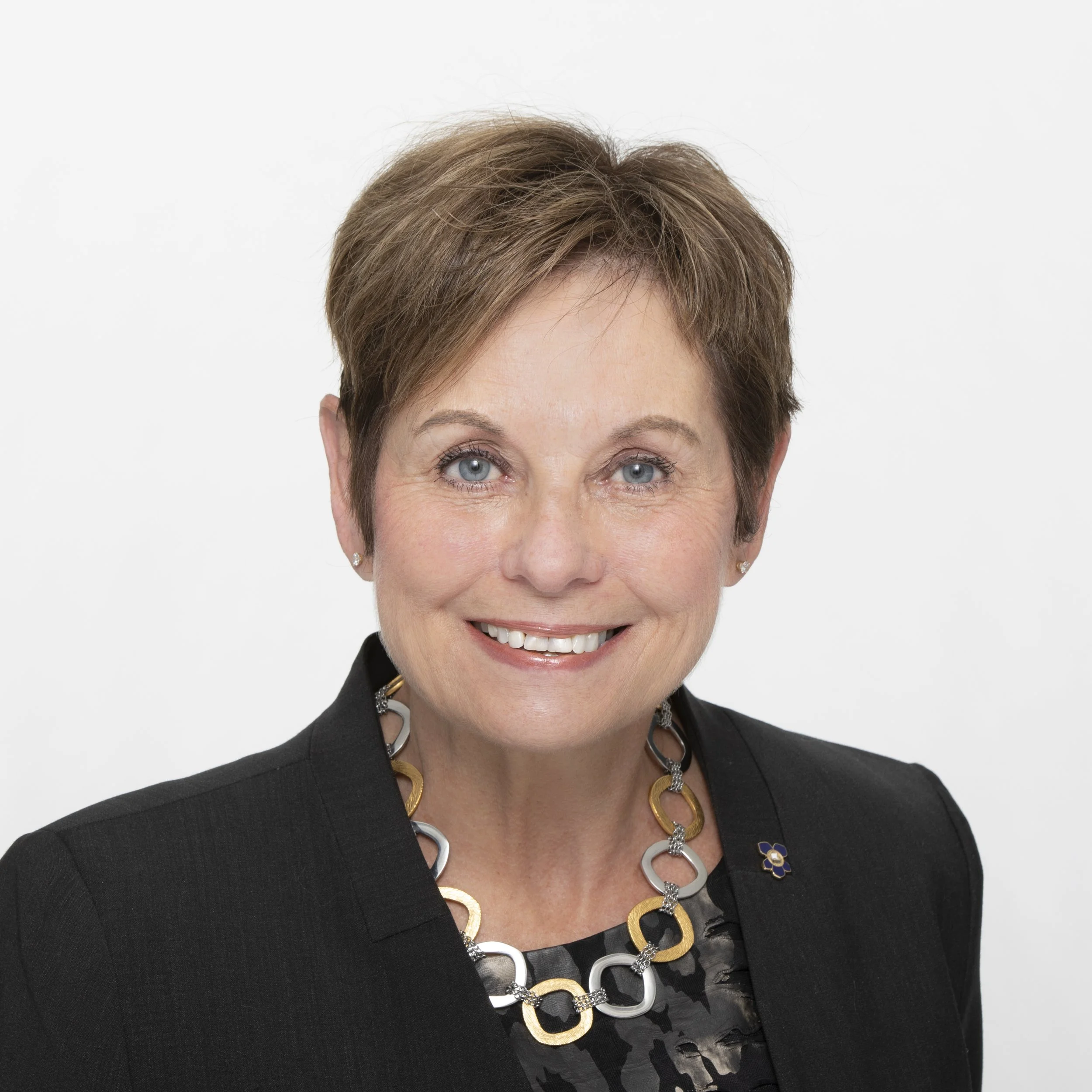 Portrait of a smiling middle-aged woman with short brown hair, wearing a black blazer, a black patterned blouse, and a multicolored necklace against a plain white background.