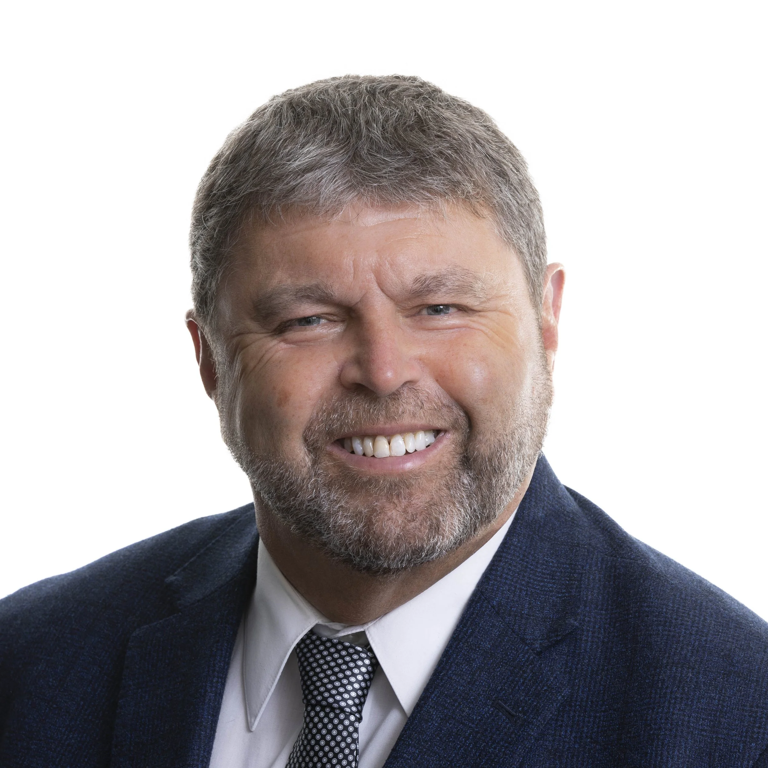 Portrait of a middle-aged man with gray hair and a beard, smiling, wearing a dark suit, white shirt, and patterned tie, against a white background.