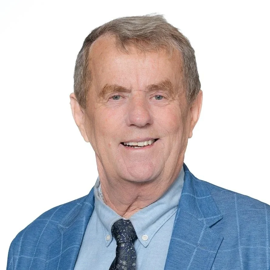 A professional portrait of an older man with glasses, gray hair, and a mustache, wearing a dark brown suit jacket, white shirt, and striped tie, smiling against a plain white background.