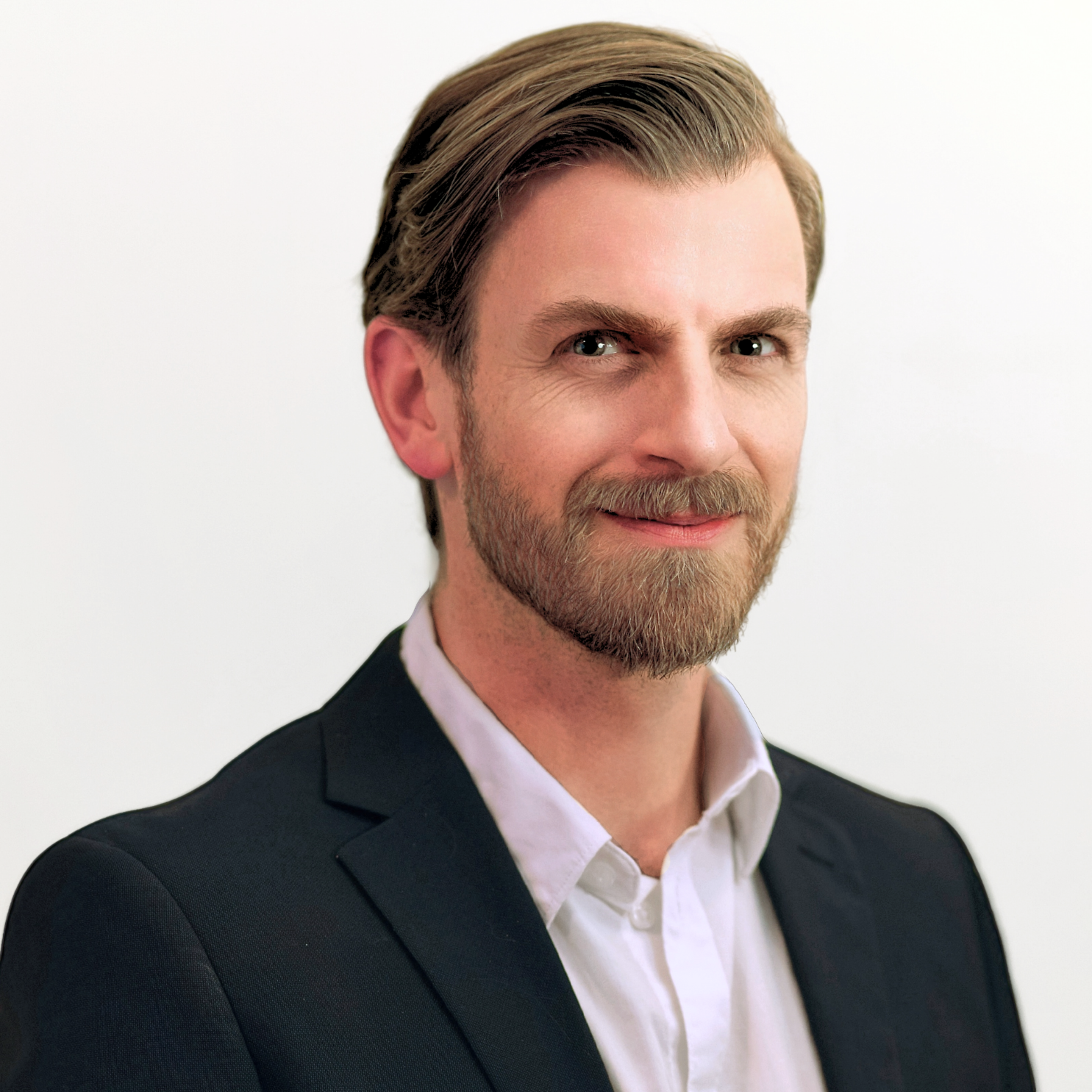 Portrait of a smiling man with light brown hair, a beard, wearing a dark suit jacket over a white collared shirt against a plain white background.