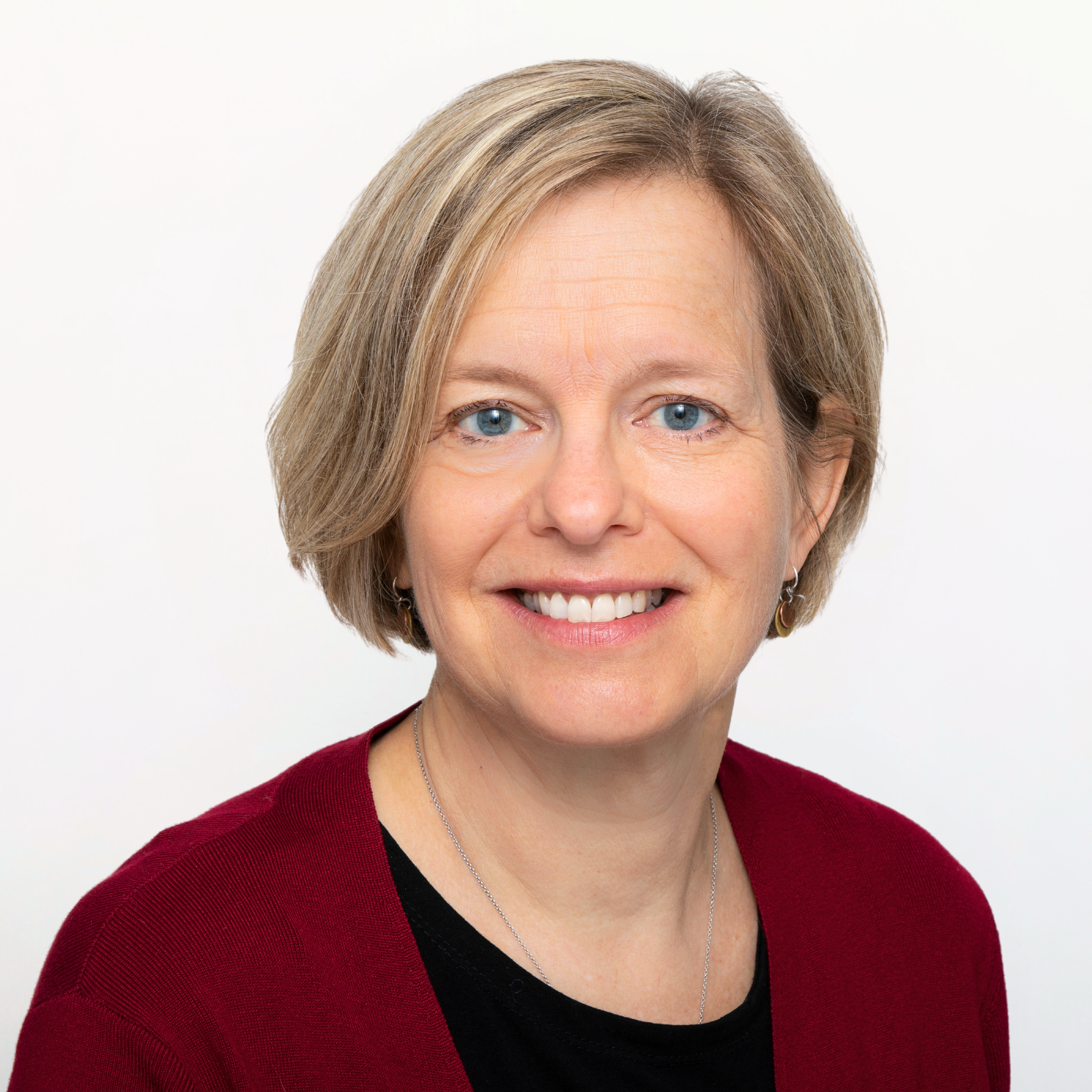 A middle-aged woman with short blonde hair, blue eyes, and a warm smile, wearing a red sweater over a black top and silver jewelry, posing against a plain white background.