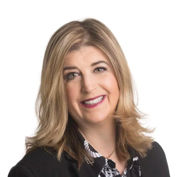 A woman with shoulder-length blonde hair, smiling, wearing a black blazer over a patterned blouse, against a white background.