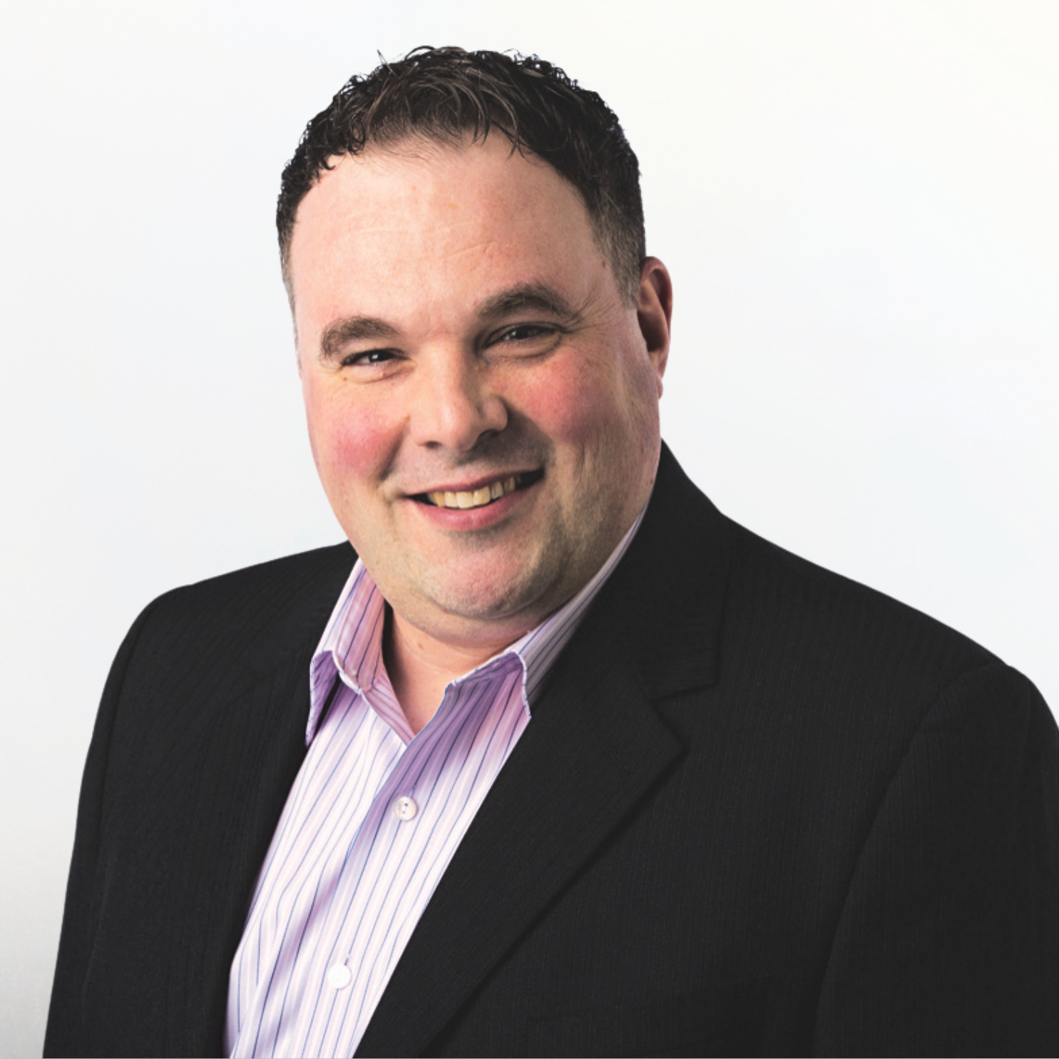 A professional headshot of a smiling man with short dark hair, wearing a black suit jacket and a pink striped dress shirt, against a plain white background.