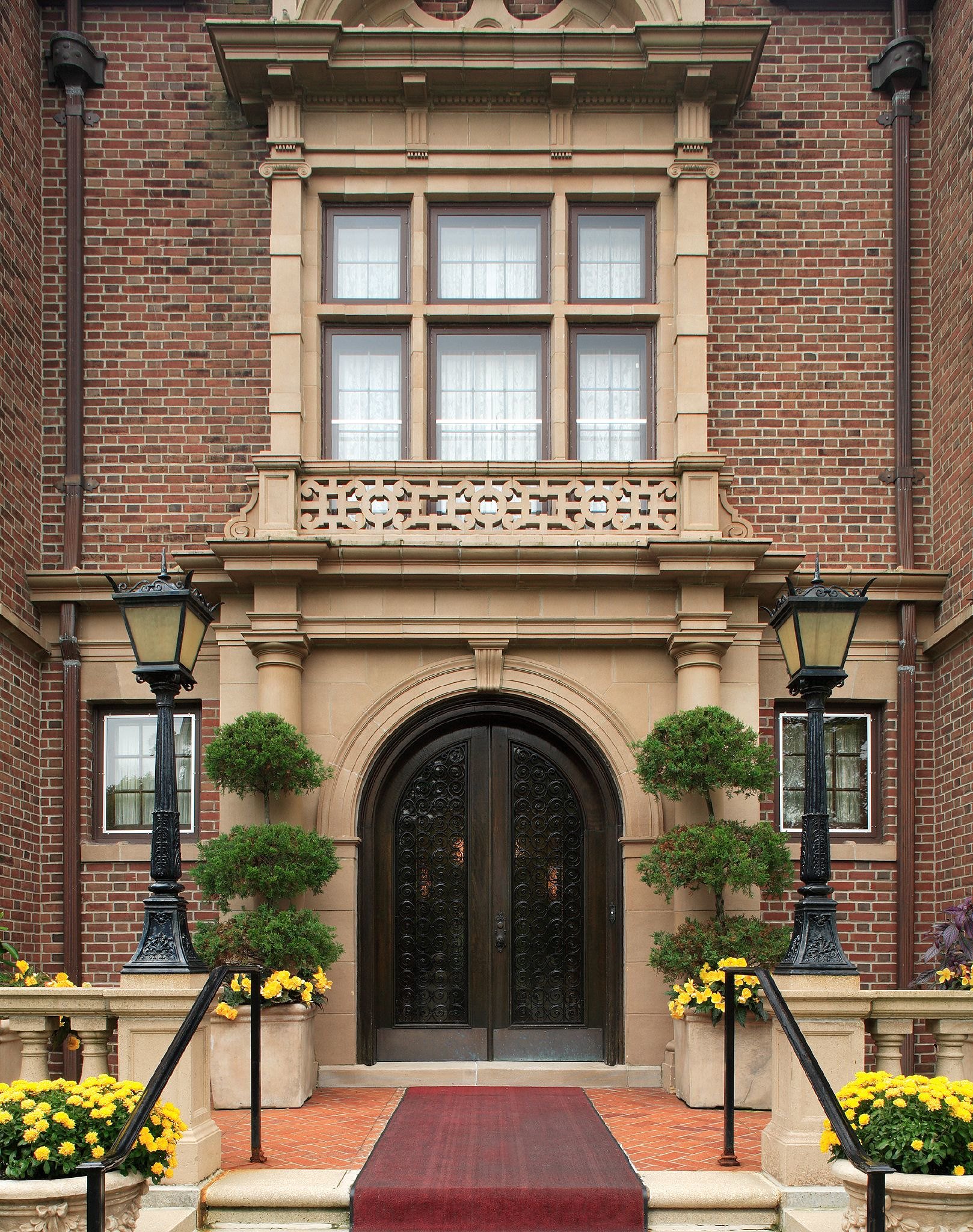 Elegant brick building entrance with arched black door, potted trees, yellow flowers, and vintage street lamps. Governor residence in Saint Paul, MN.