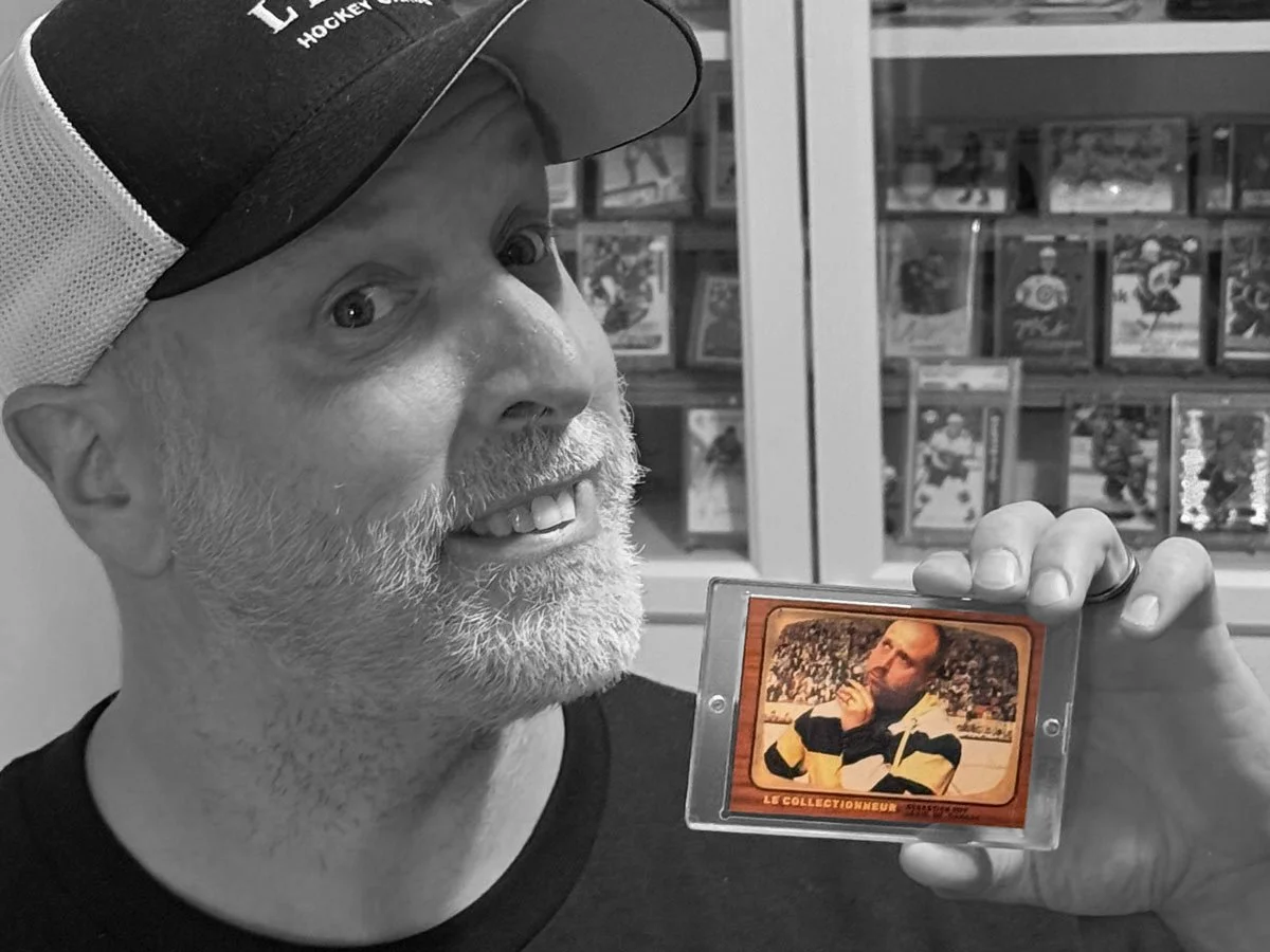 Une personne souriante portant une casquette, tenant une carte de collection vintage en couleur, avec des étagères remplies de boîtes de jeux ou de cartes en arrière-plan.