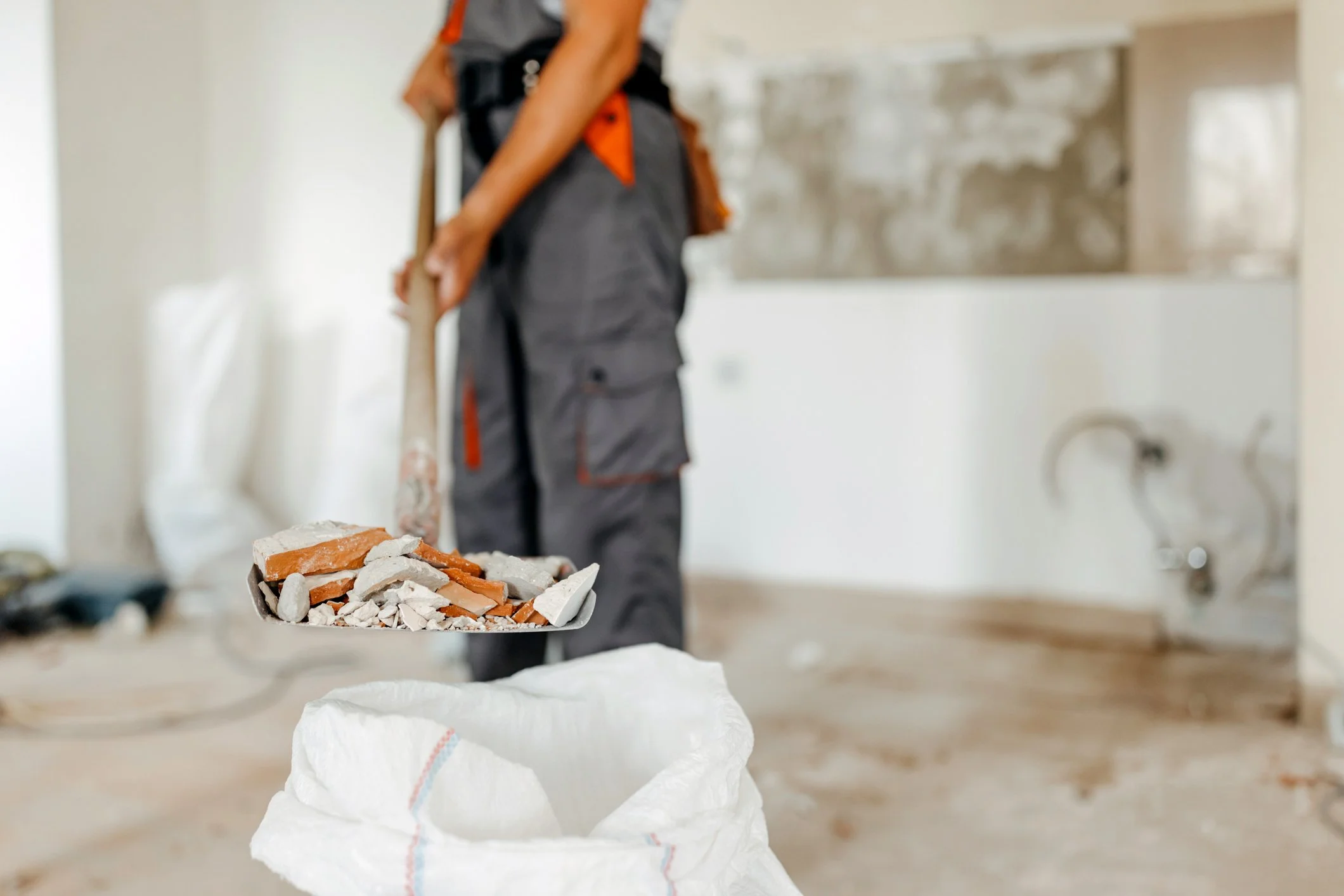 Een persoon staat in een keuken of bouwruimte met een schep vol afgebroken bakstenen en ander puin, klaar om te verwijderen.