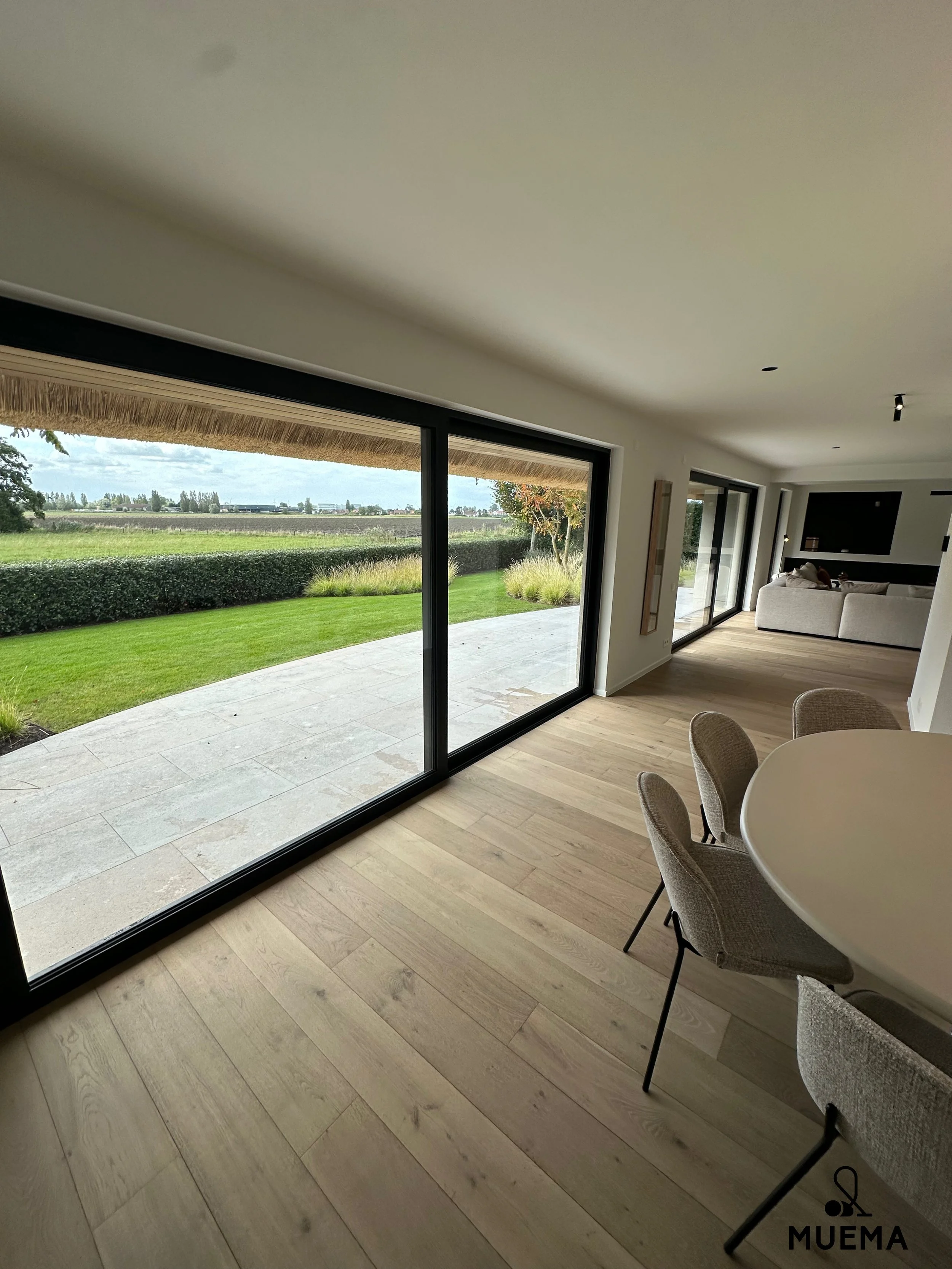 Een moderne woonkamer met grote glazen schuifdeuren die uitkijken op een tuin met gras, struiken en een horizon met bomen.