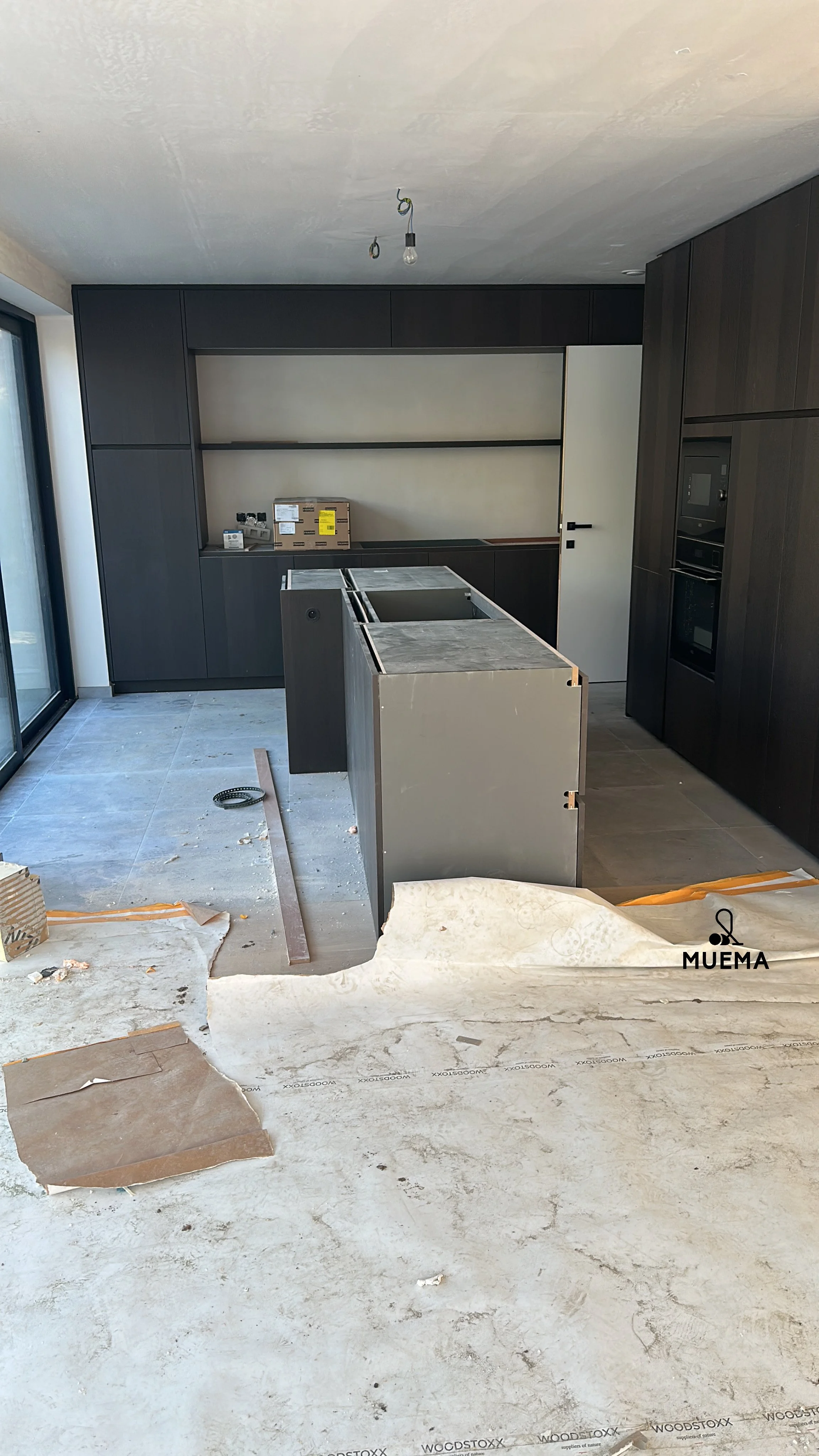 Binnenkant van een keuken onder renovatie met nog te plaatsen keukeneiland, zwarte kasten en apparaten, en een plafond met blootliggende lampdraden.