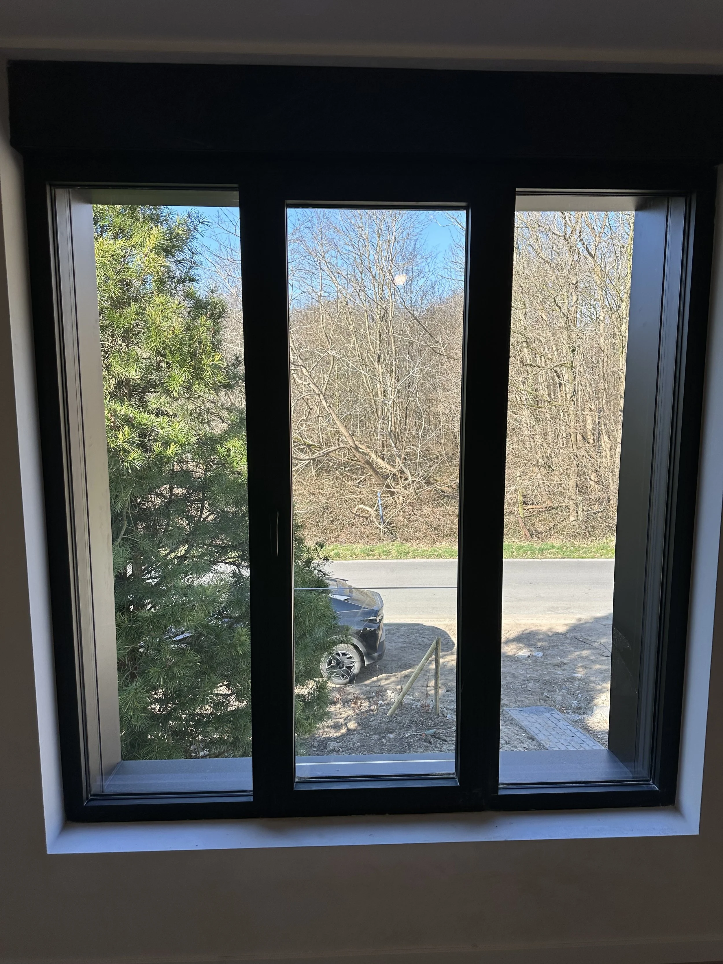 Uitzicht door een groot raam met drie panelen, met bomen en een auto buiten.