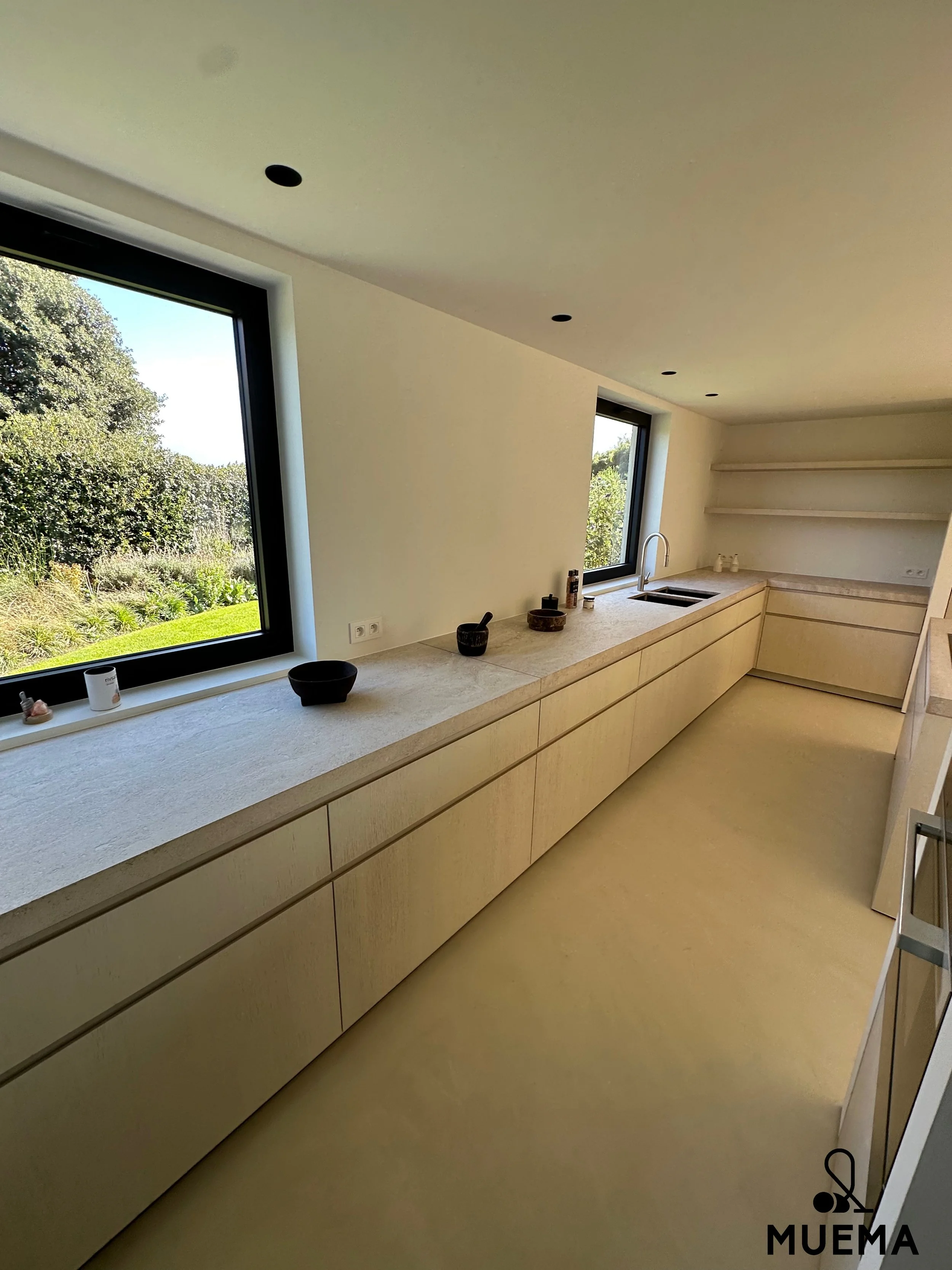 Lichte moderne keuken met lange witte kasten onder twee grote ramen die uitzicht bieden op een groene tuin.