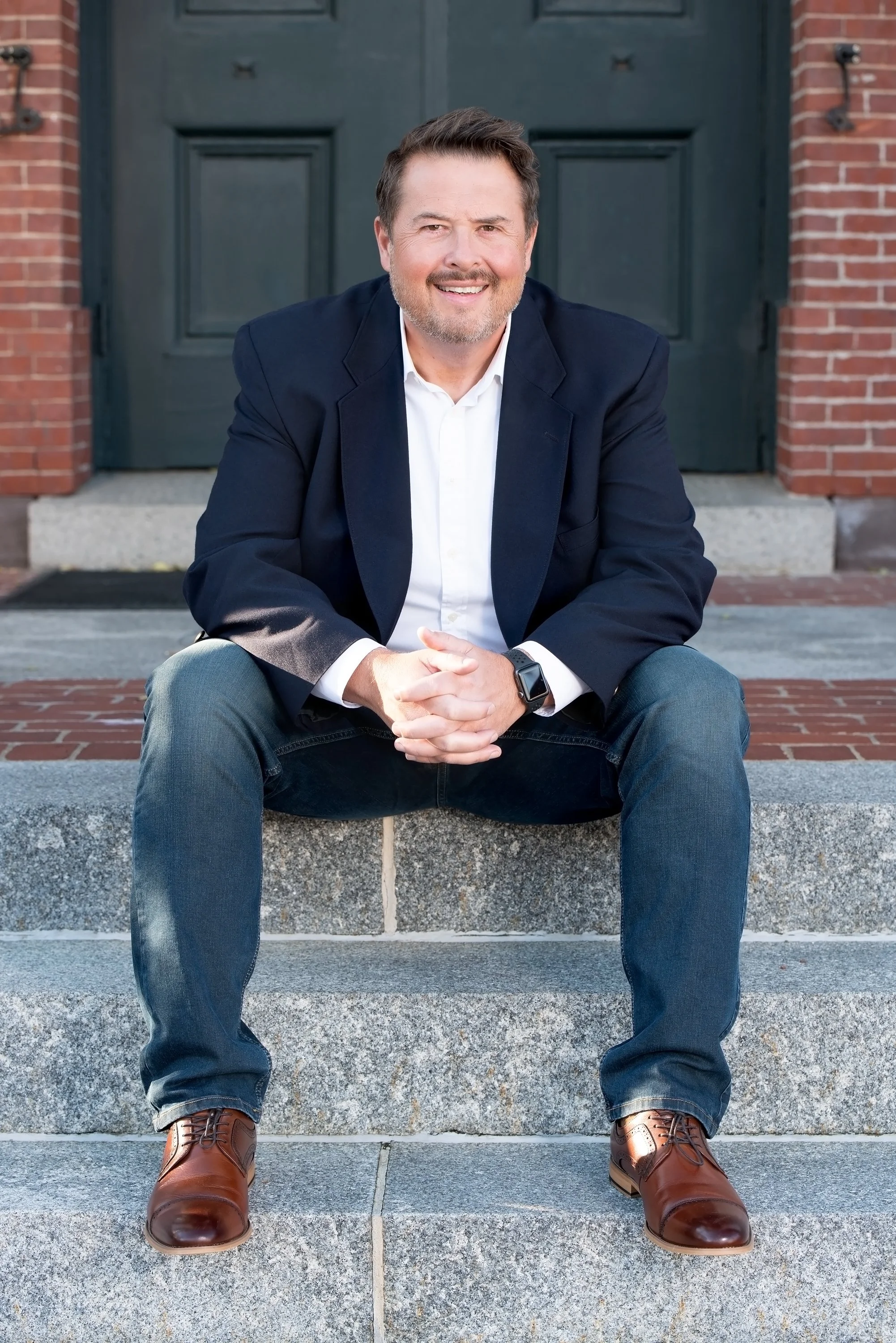 A man sitting on stone steps in front of a dark green door, smiling and wearing a navy blazer, white shirt, blue jeans, brown shoes, and a smartwatch.