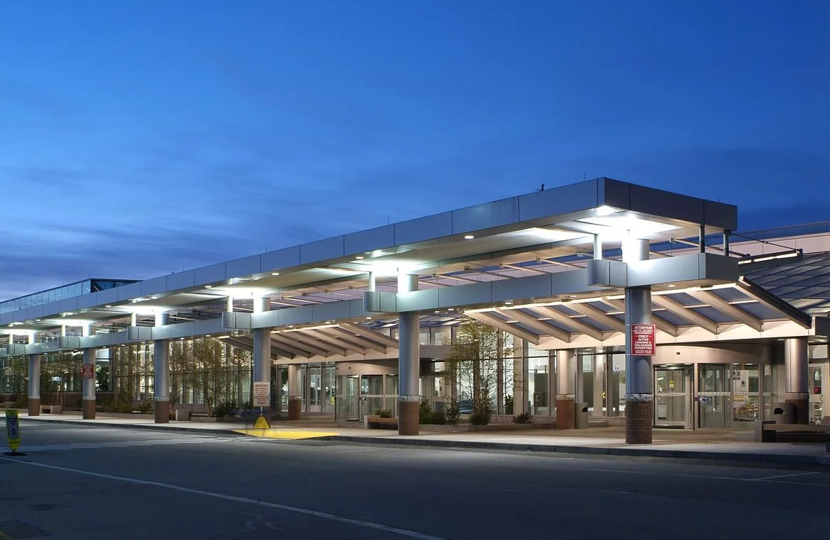 Modern hospital entrance with glass doors, covered drop-off area, and bright lighting at dusk.