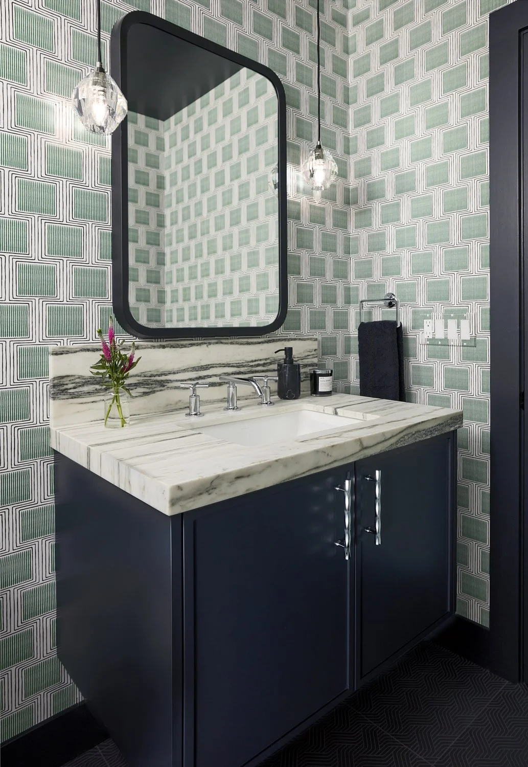 A bathroom vanity with a white marble countertop, dark blue cabinet, and a rectangular mirror with a black frame. The wall behind has green geometric patterned wallpaper, and there are two glass pendant lights hanging from above. The vanity has a sma