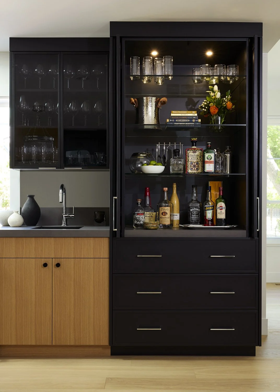 A black kitchen display cabinet filled with glassware, bottles, and decorative items, adjacent to a wooden kitchen counter with a sink and vase.