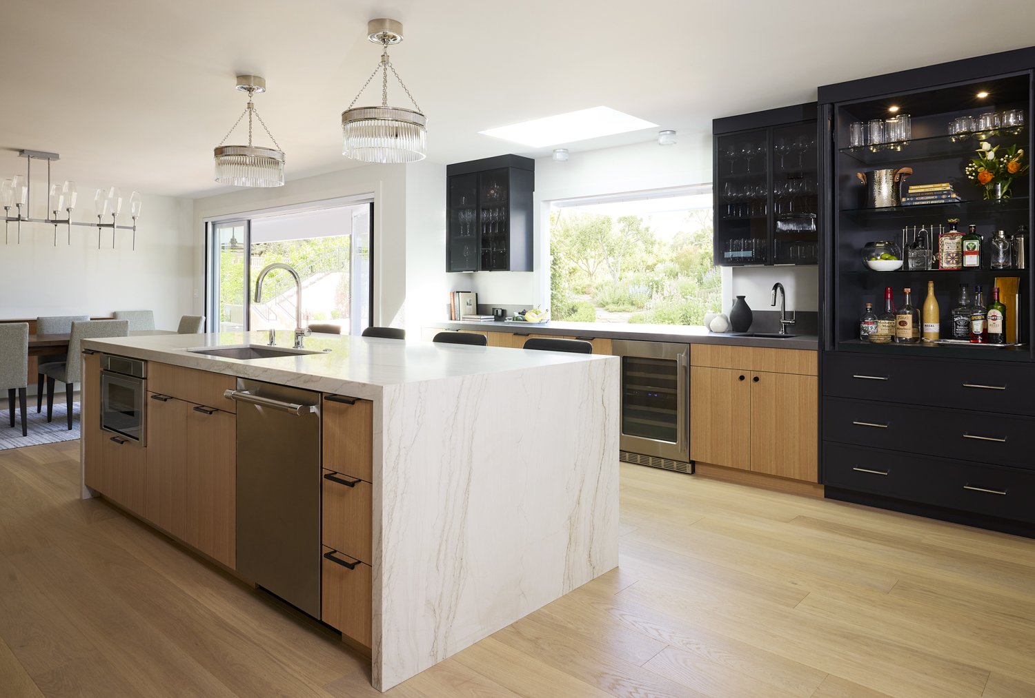 Modern kitchen with white marble island, black and wood cabinets, bar area with liquor bottles, and large windows showing greenery outside.