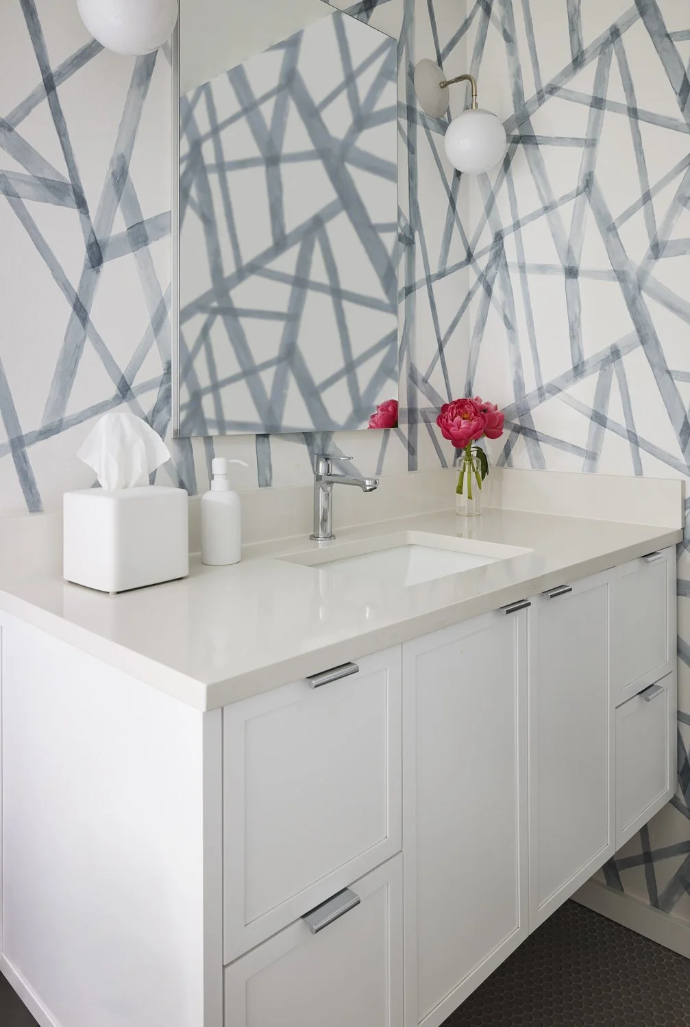 Bathroom vanity with a white counter, a sink, a mirror, a decorative vase with pink flowers, a tissue box, a soap dispenser, and wall-mounted light fixtures, with geometric patterned wallpaper.