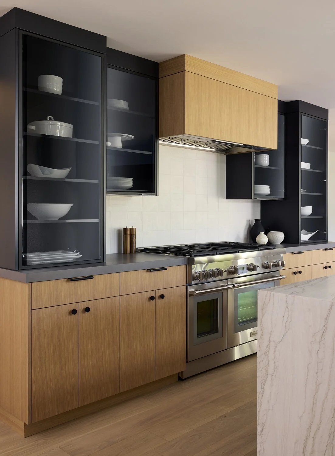 Modern kitchen with wooden cabinets, black glass-front upper cabinets, stainless steel oven, and a marble kitchen island.