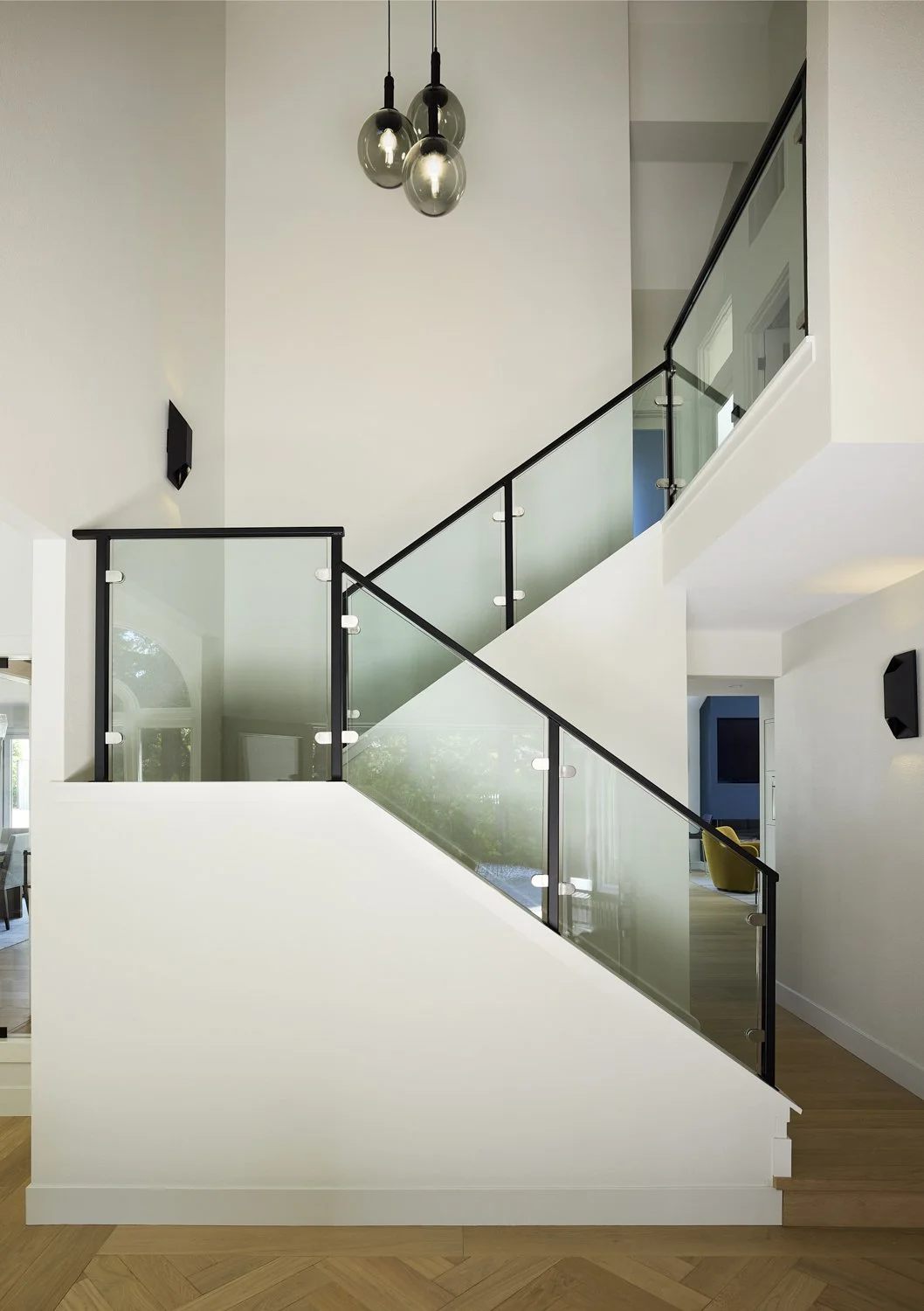 Interior of a modern home showing a staircase with glass and black metal railing, hanging pendant lights, and white walls.