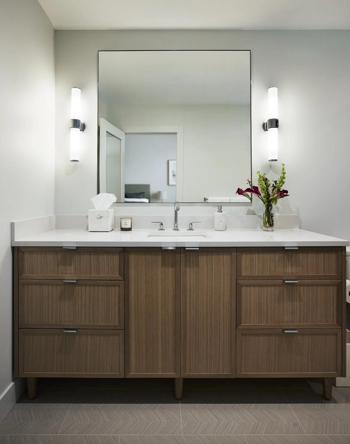 A bathroom vanity with a large mirror, two modern wall-mounted lights, a white countertop, a tissue box, a candle, and a vase with flowers.