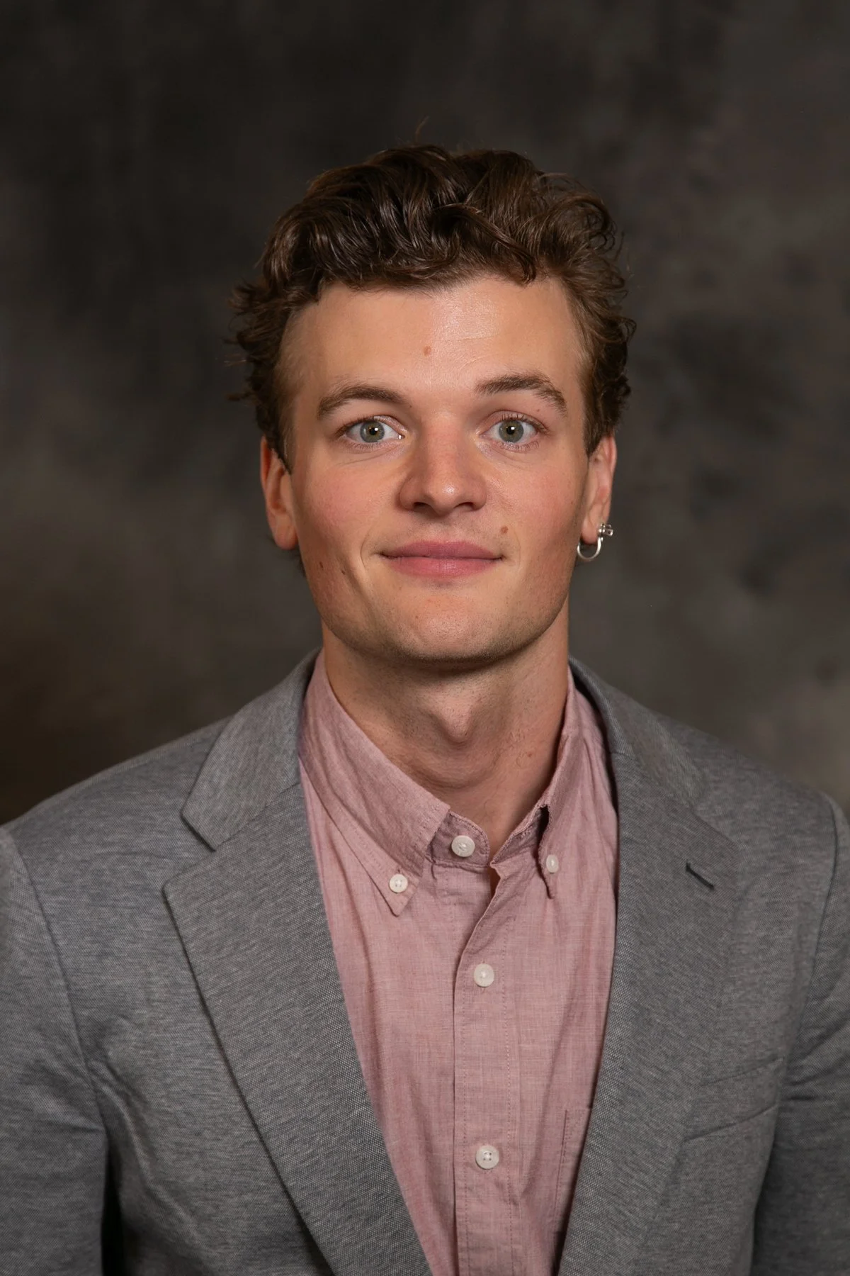 A young man with short curly brown hair, light skin, and blue eyes, wearing a gray blazer over a pink button-up shirt, against a dark background.