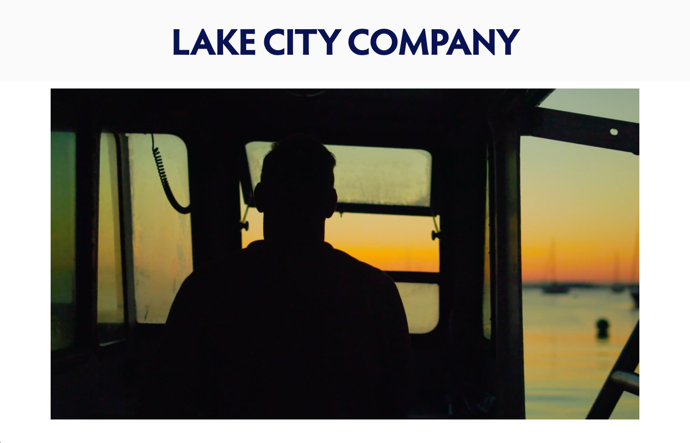 Silhouette of a person sitting inside a boat cockpit at sunrise, with water and boats in the distance.