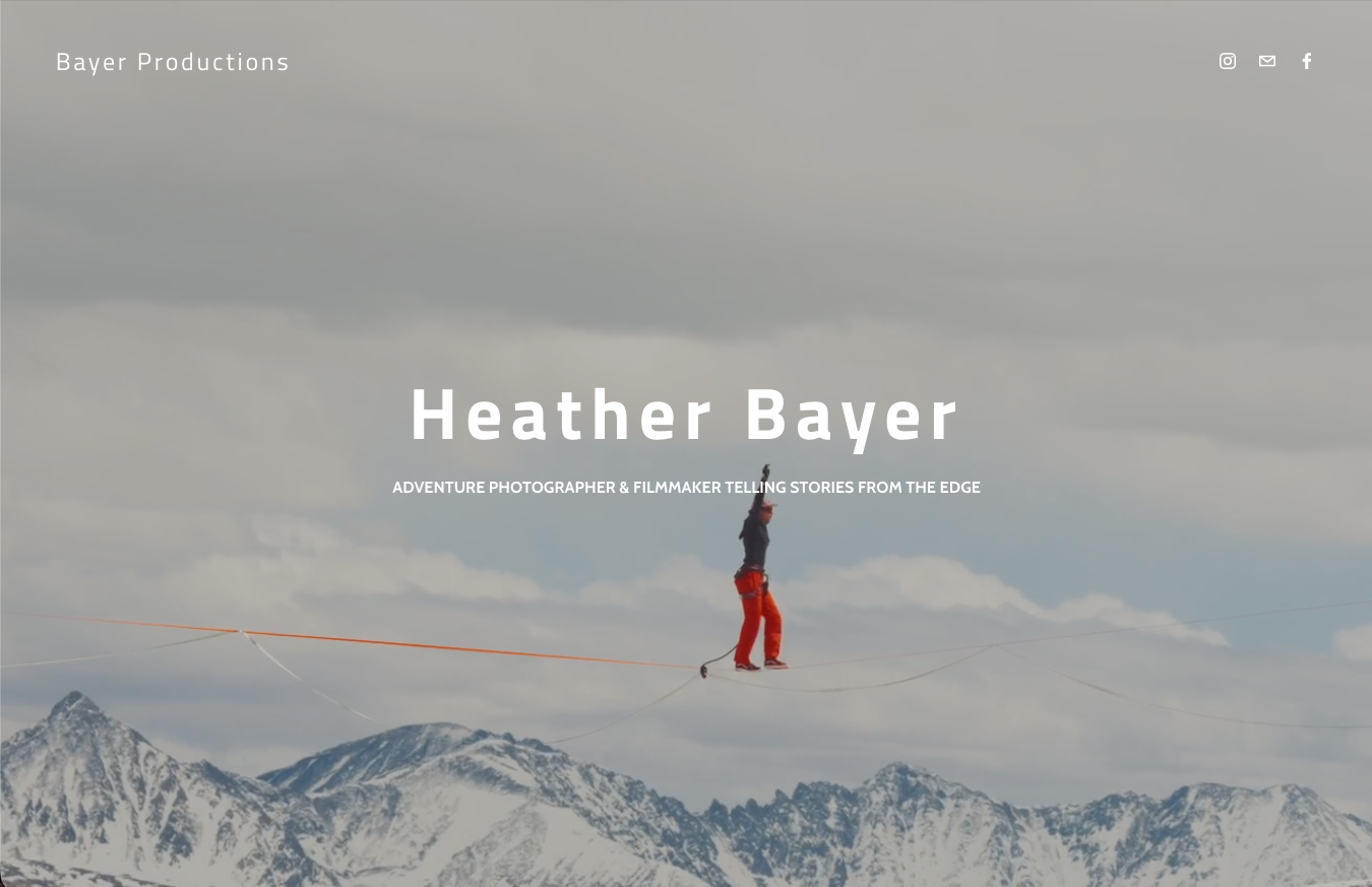 A person dressed in red pants and a dark jacket walking on a tightrope stretched across snow-covered mountain peaks under a cloudy sky.
