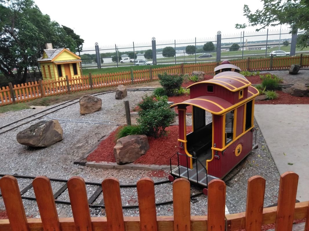 Miniature garden train set with tracks, small red train car, and yellow house inside fenced area