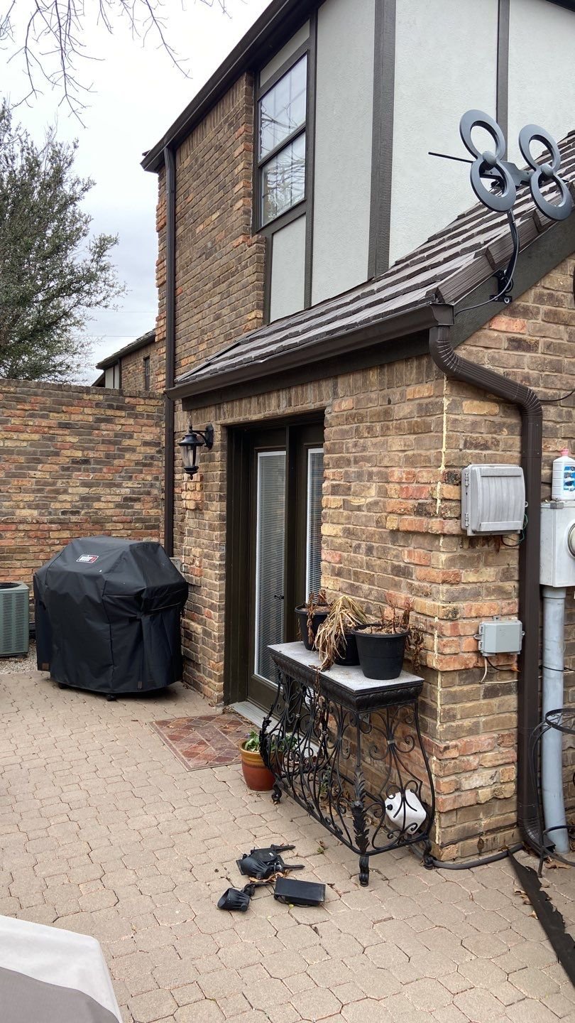 Backyard patio with a brick exterior, covered grill, decorative black metal table, potted plants, and an antenna attached to the roof.