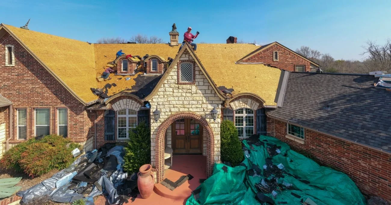 House with roof under repair, workers on roof, roofing materials on ground, and house surrounded by trees.
