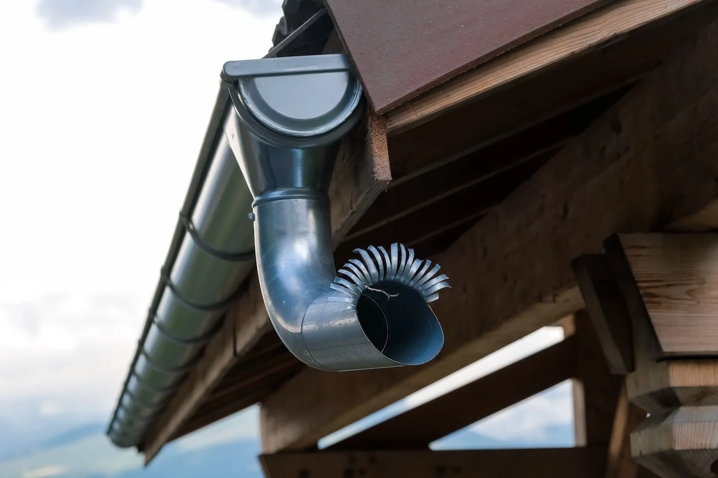 Close-up of a metal gutter downspout attached to a wooden roof structure, with a background of cloudy sky and distant landscape.