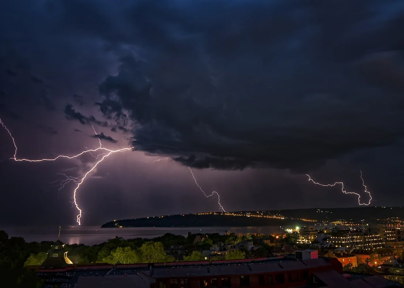 Dark stormy sky with multiple lightning strikes over a cityscape near a body of water at night.