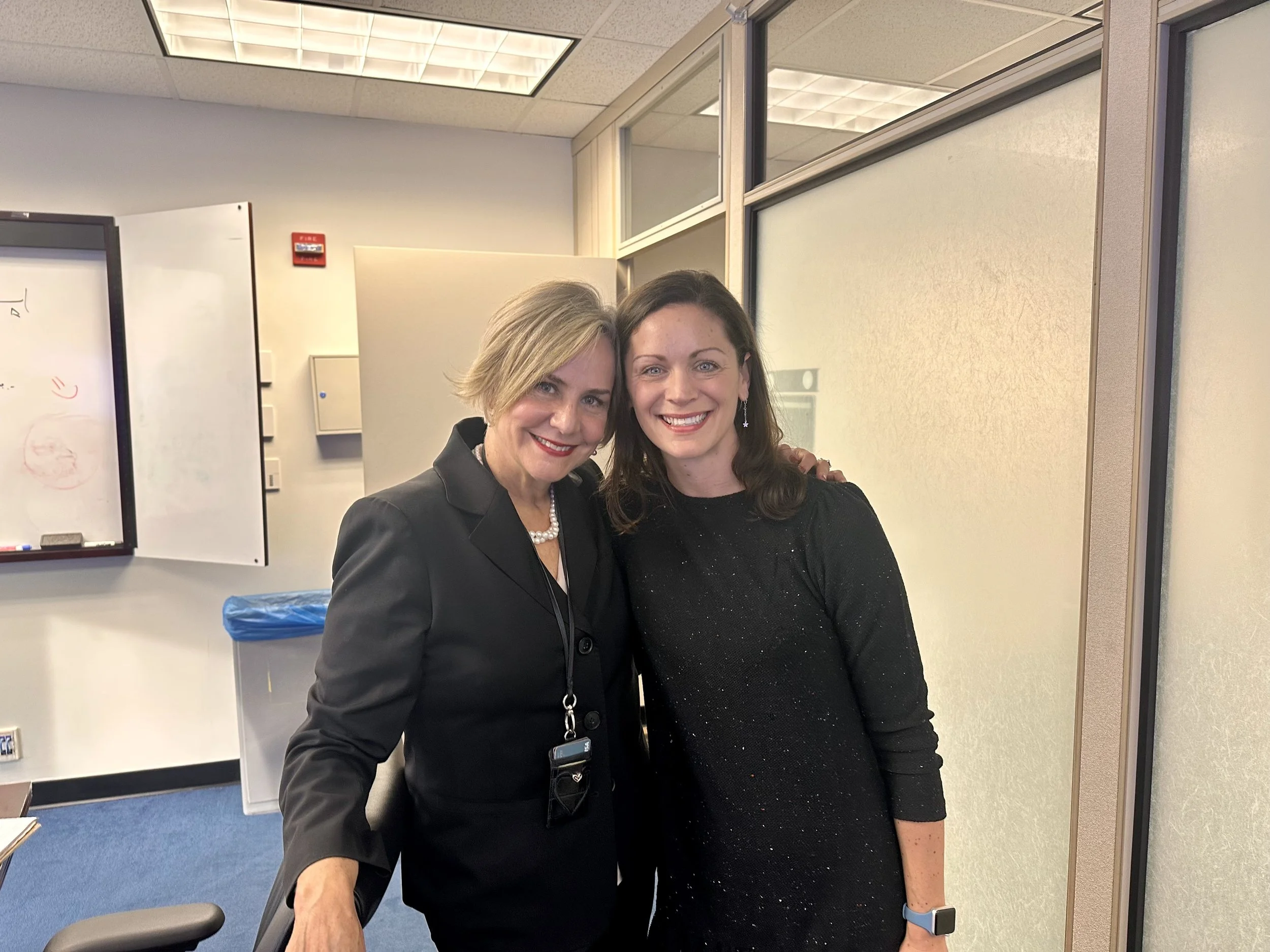 Two women smiling and standing together in an office, with one woman in a black suit and the other in a black sparkly dress.