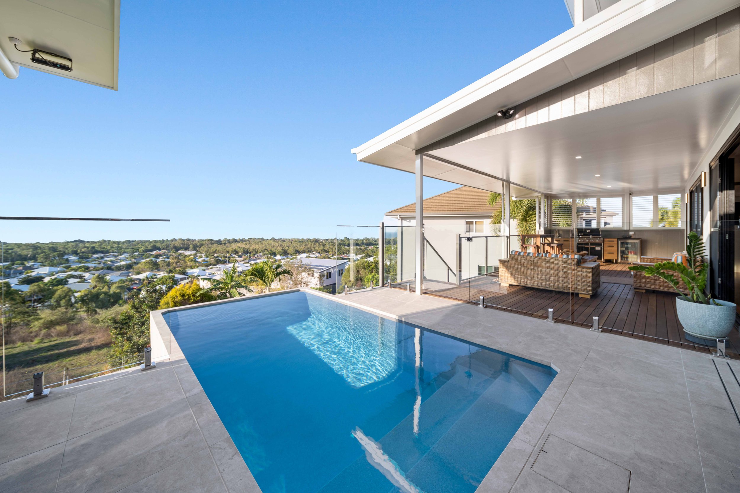 Modern outdoor balcony with a small rectangular swimming pool, sitting area with wicker sofas, and panoramic view of a suburban neighborhood with trees and houses under a clear blue sky.
