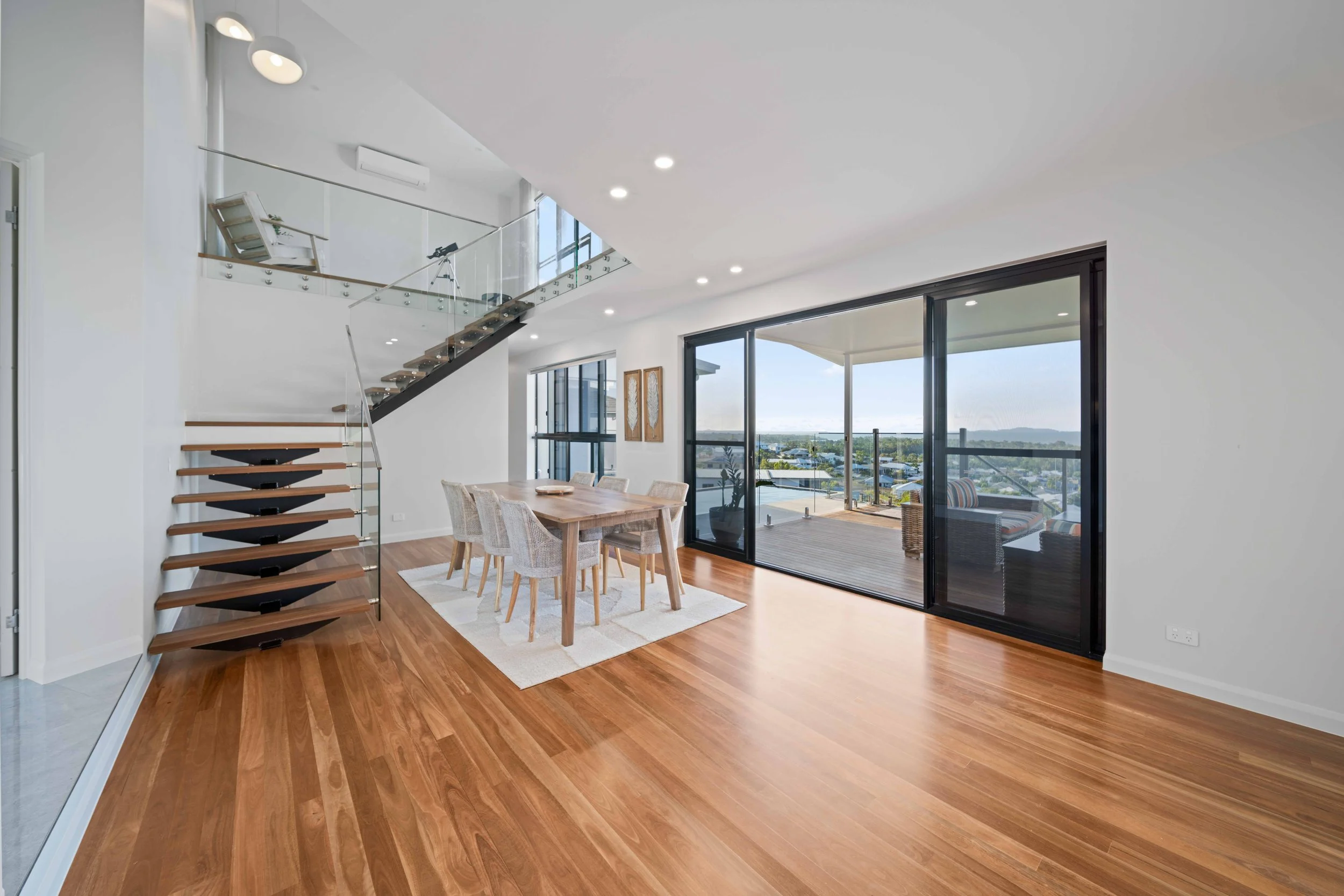 Open living and dining area with wooden floors, a dining table with eight chairs, large sliding glass doors leading to a balcony, and staircase with glass railing.
