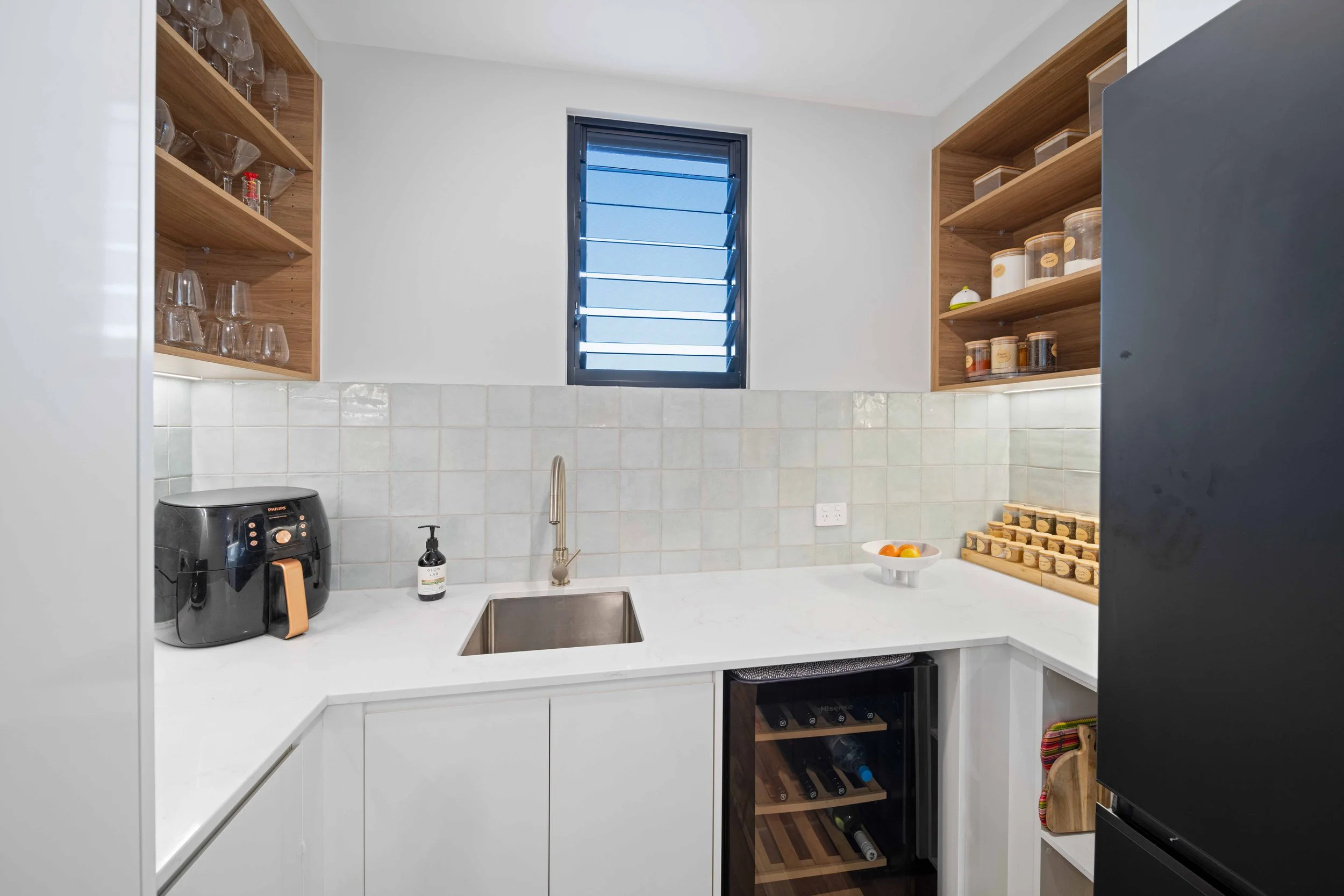 Modern kitchen with white countertops, wooden open shelves holding glassware and containers, a small window, a black air fryer, a soap dispenser, a stainless steel sink with a high faucet, a bowl of oranges, a wine refrigerator, and a black refrigera