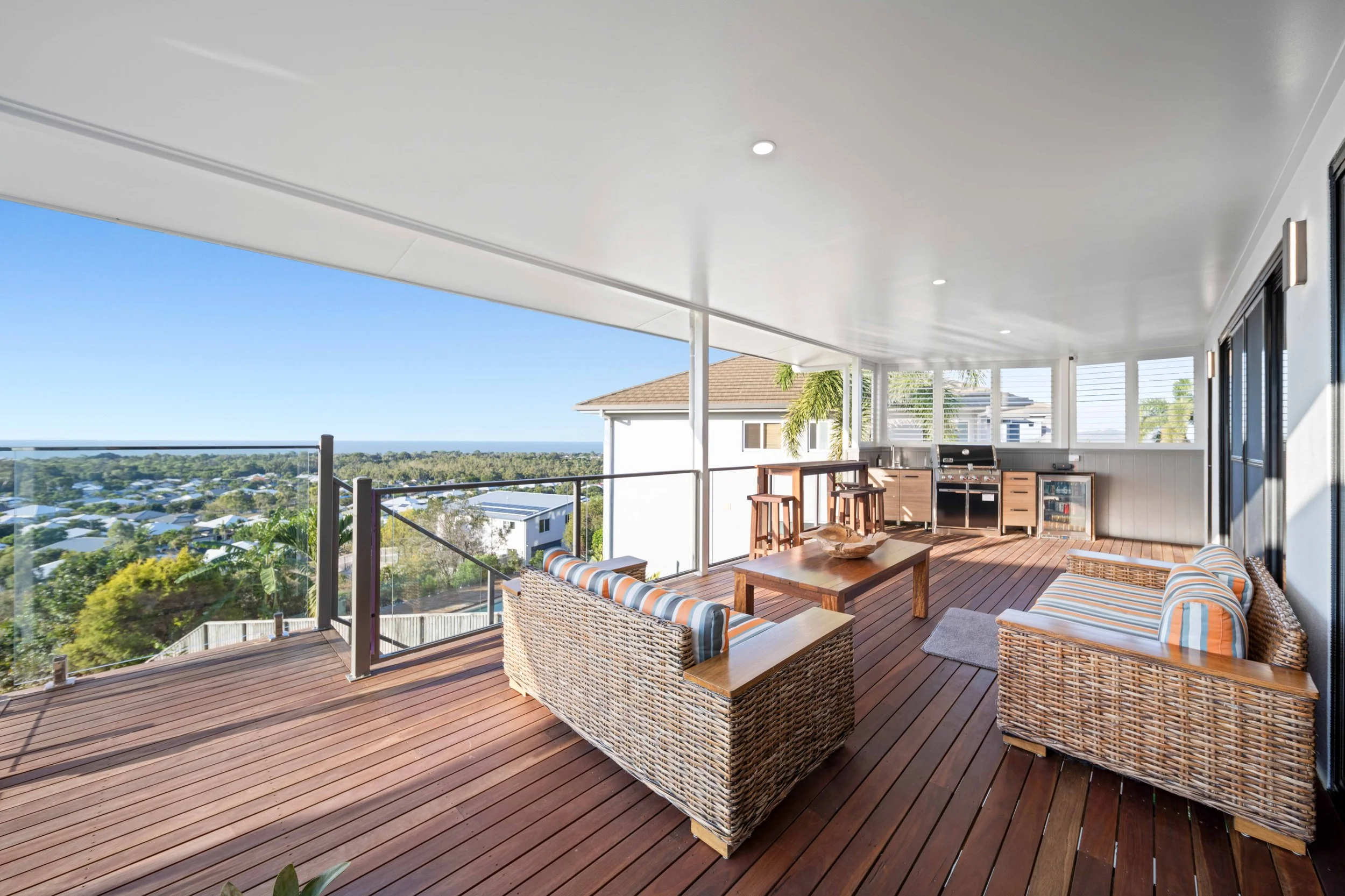 Covered outdoor balcony with wicker furniture, wooden table, barbecue grill, and ocean views.