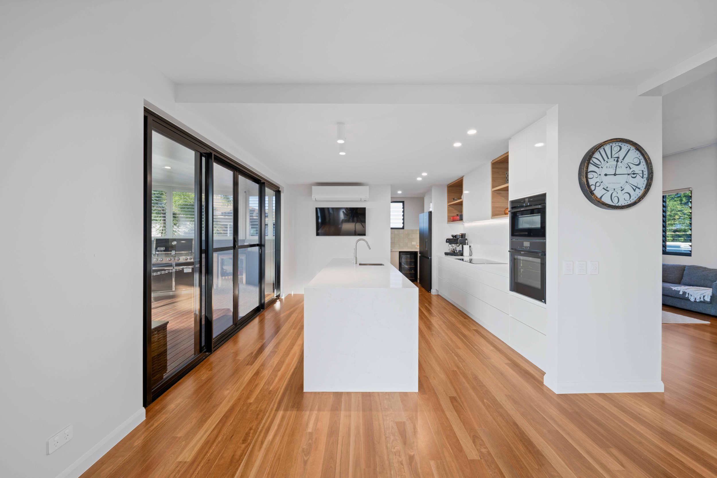 Modern kitchen with white cabinets, wooden open shelves, stainless steel appliances, a large island with a sink, hardwood flooring, sliding glass doors, and a wall clock.