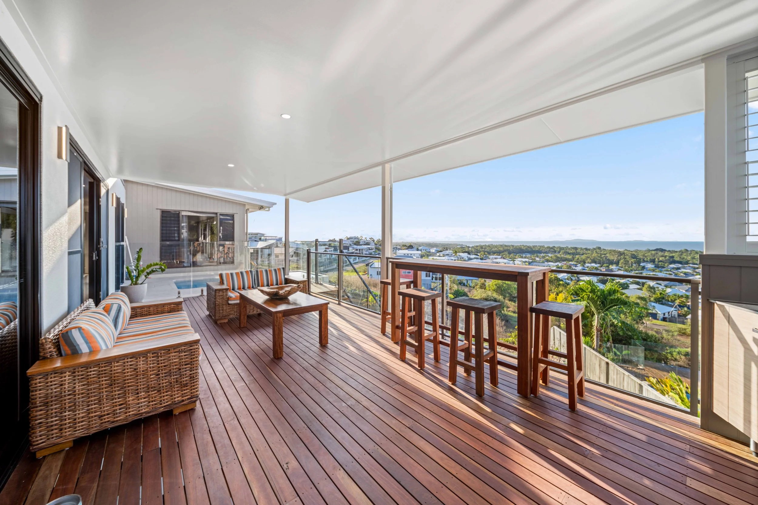Spacious outdoor balcony with wicker and wooden furniture, overlooking a scenic view of houses, trees, and the horizon on a sunny day.