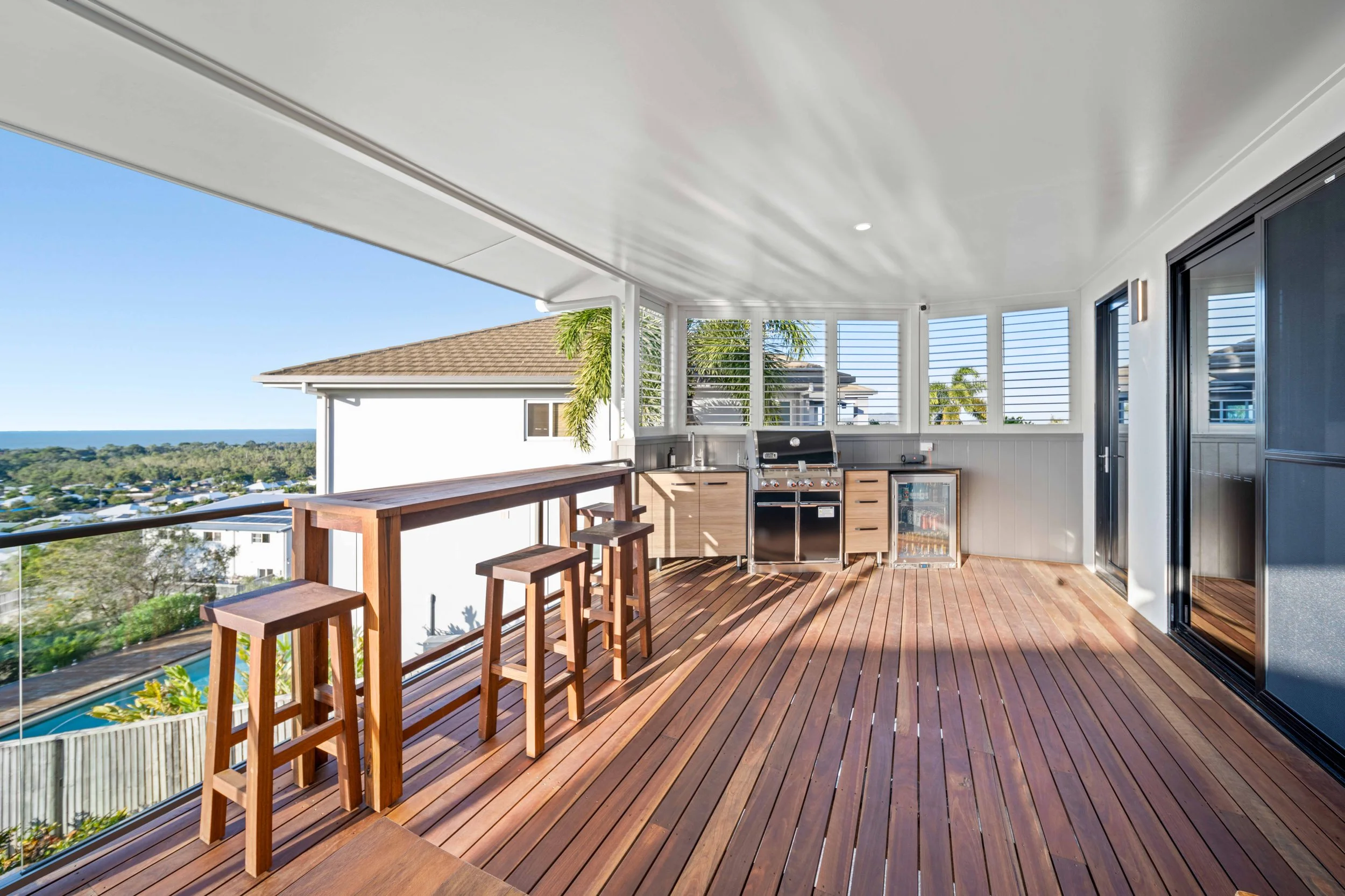 Covered balcony with wooden flooring, outdoor kitchen with grill, mini fridge, and cabinets, bar-height wooden table with stools, and views of houses, trees, and the ocean in the distance.