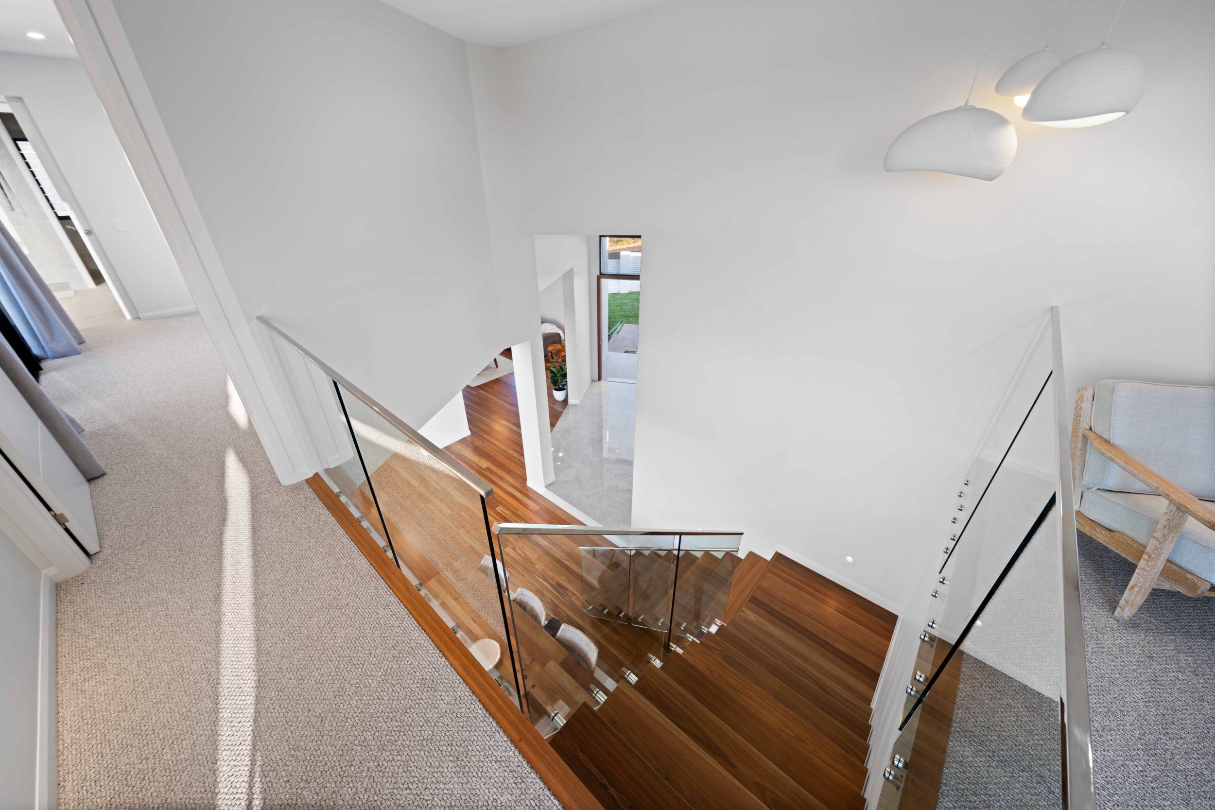 Interior view of a modern staircase with wooden steps and glass railing, overlooking the ground floor of a house with hardwood flooring and white walls.
