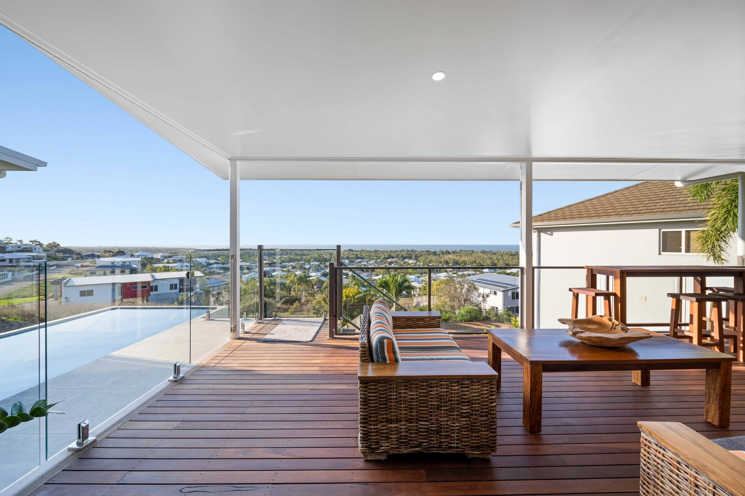 Covered outdoor balcony with wooden floor, glass railing, wooden dining table, wicker sofa with striped cushions, and barstools, overlooking a suburban neighborhood with a view of trees and the ocean in the distance.