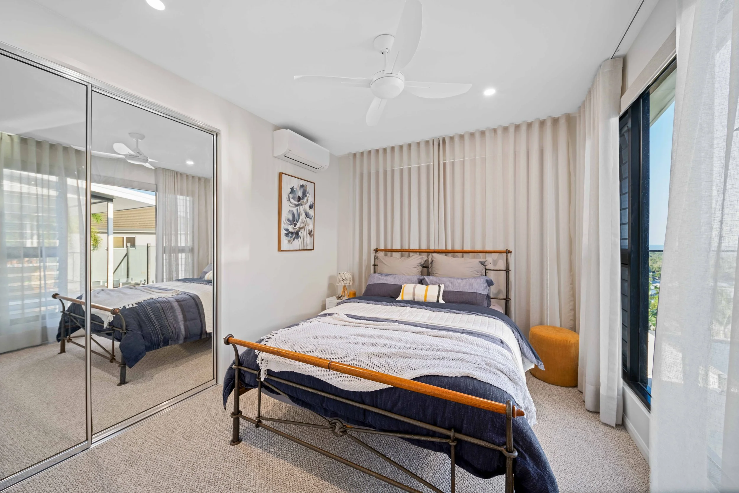 Modern bedroom with a metal bed frame, bedding, floral wall art, large closet with mirrored doors, ceiling fan, air conditioning unit, large glass window with curtains, and a small yellow ottoman.