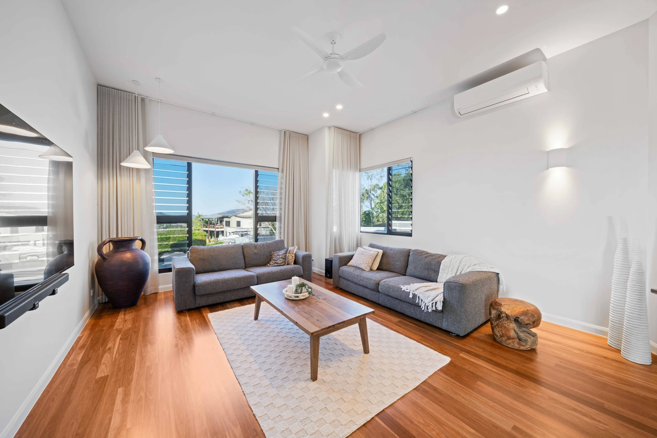 Bright living room with wooden floors, two grey sofas, a wooden coffee table, large windows with curtains, a large projector screen on the wall, a ceiling fan, an air conditioning unit, a decorative wooden stool, a large ceramic vase, and tall white 