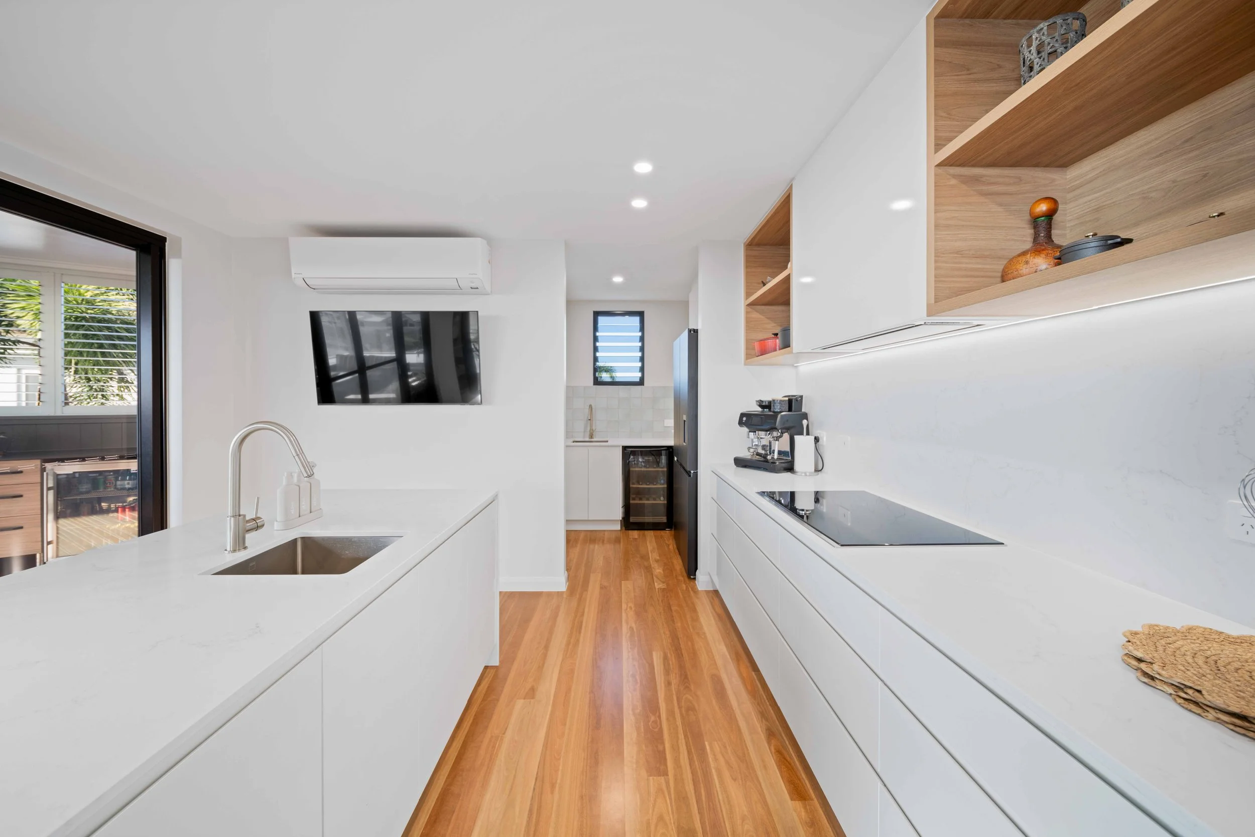 Modern kitchen with white cabinets, wooden open shelves, a black refrigerator, coffee maker, stovetop, and a wall-mounted TV. The floor is hardwood, and there is a sliding door leading outside.