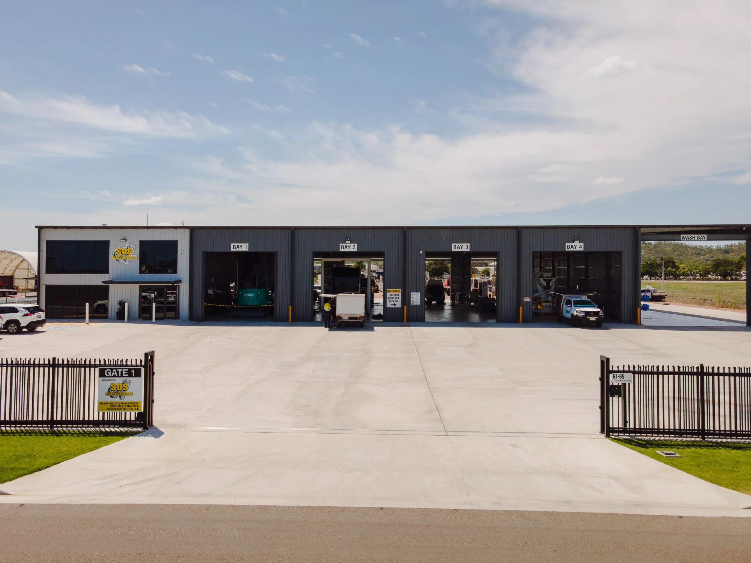A commercial vehicle wash station with five bays, labeled Bay 1 through Bay 4 and Wash Bay, including a gate with a sign for SOS vehicle wash service, parked cars, and equipment inside the bays.