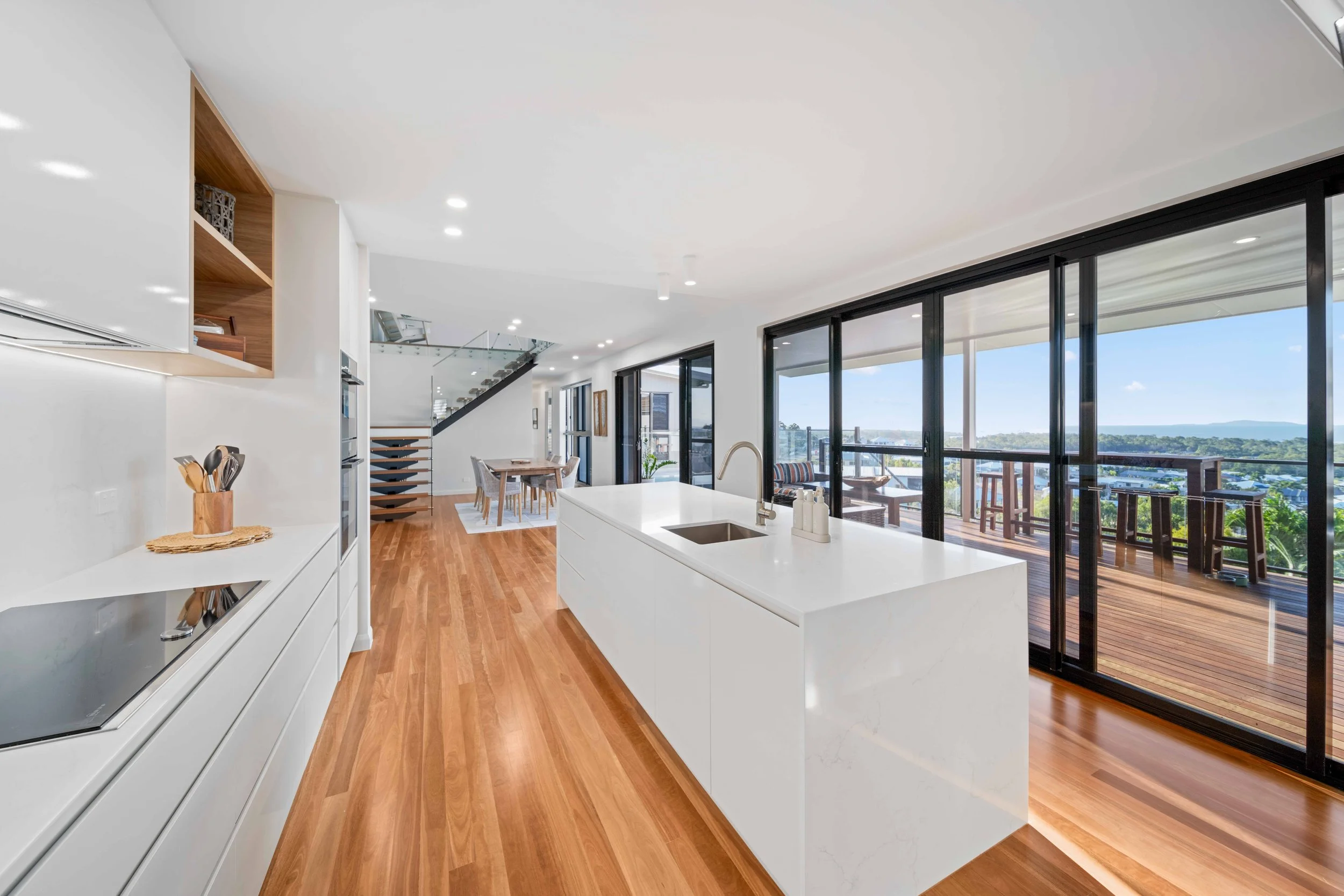 Modern kitchen with white island and wooden flooring, adjacent to balcony with outdoor seating and scenic view.