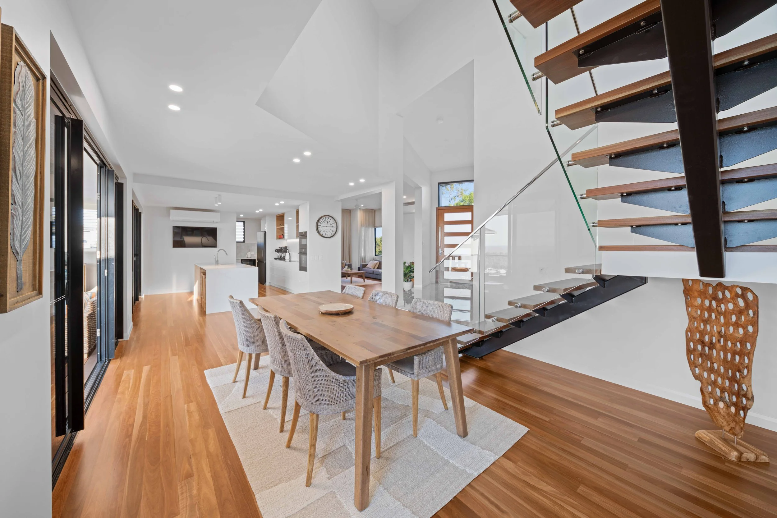 Open-concept living and dining area with a wooden dining table, six chairs, staircase with glass railing, and kitchen in the background.