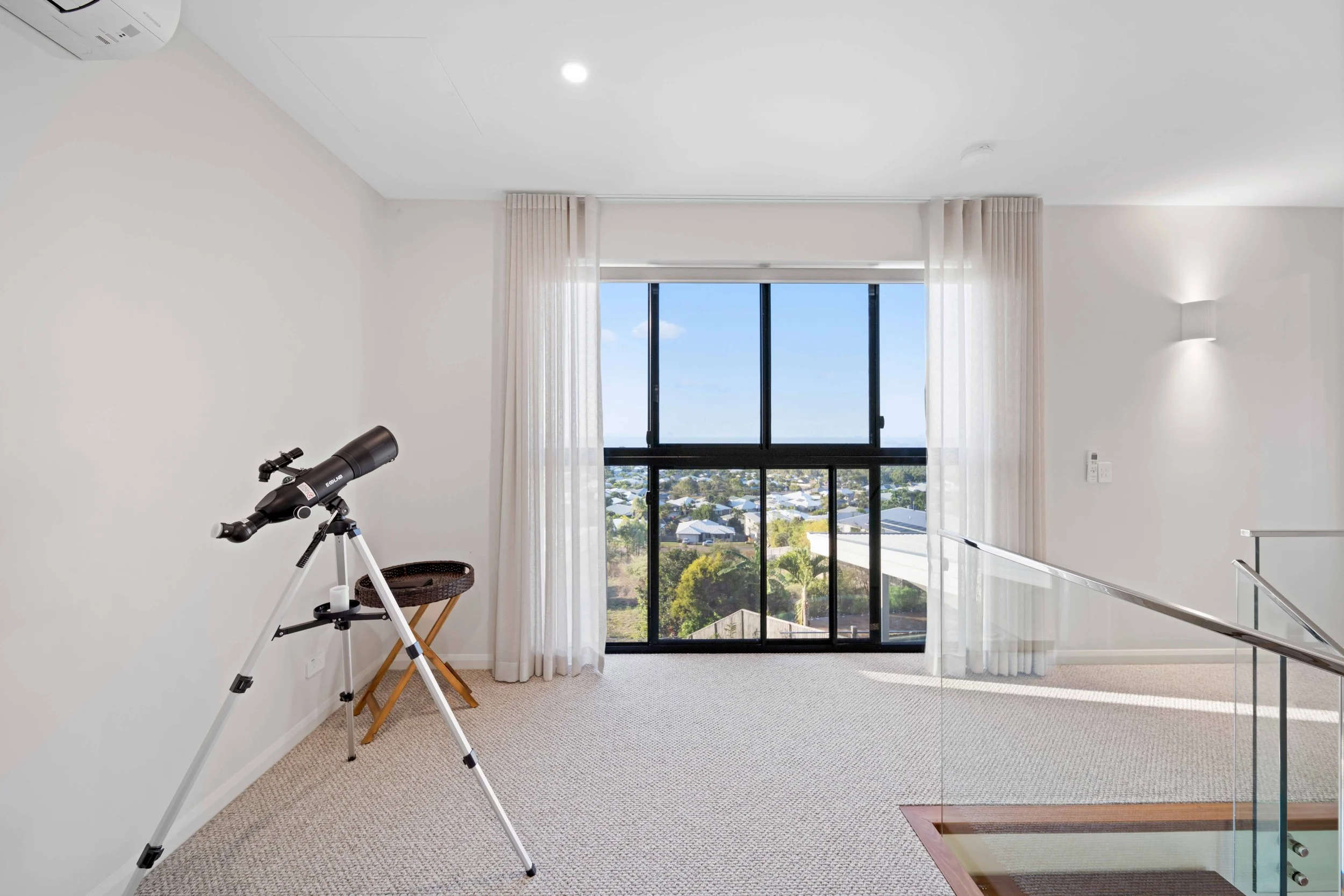Room with a telescope on a tripod near the wall, a window with sheer curtains, and a view of a neighborhood outside.
